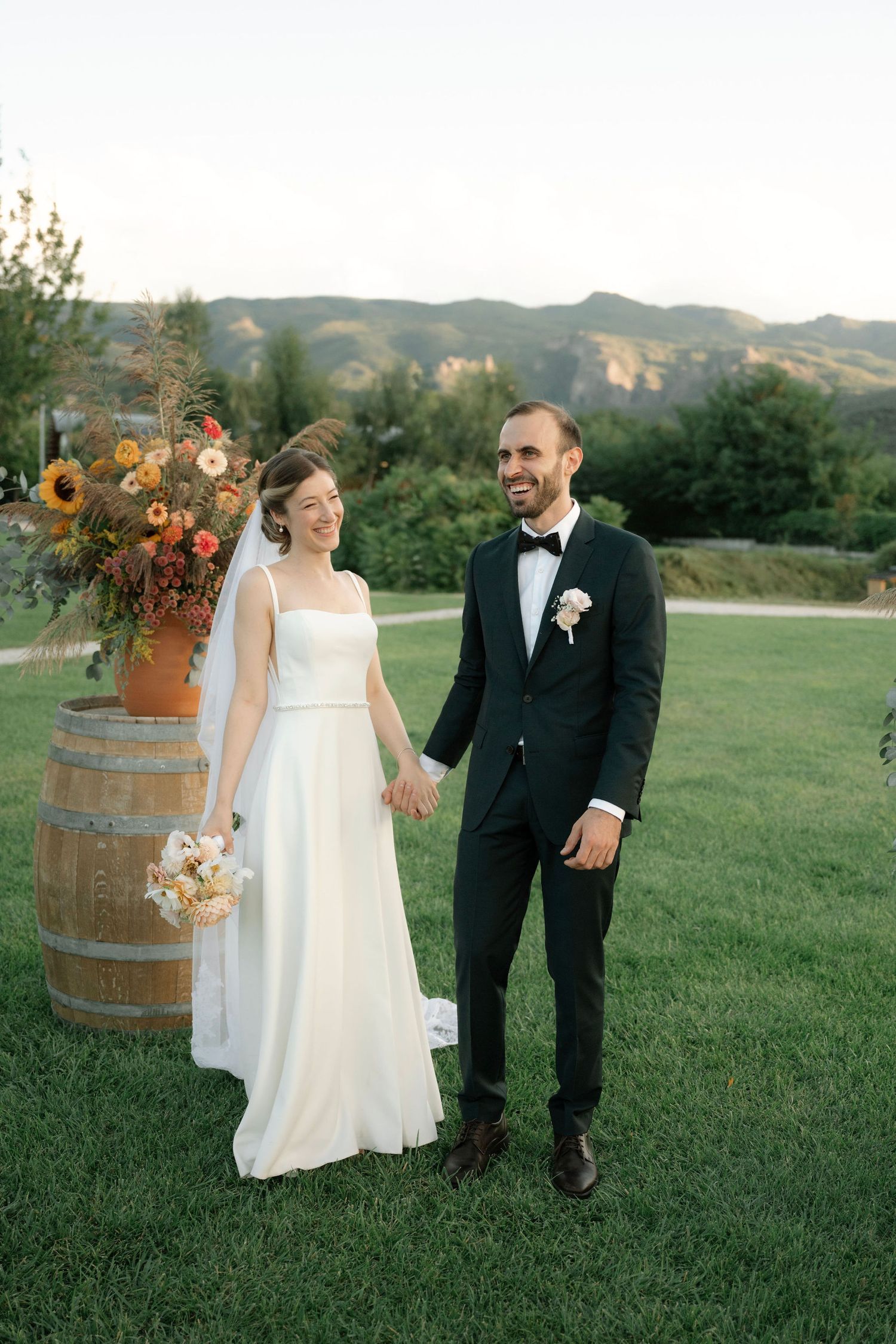 A bride and groom walk hand in hand across a lush green lawn with rolling hills and wine barrels in the background.