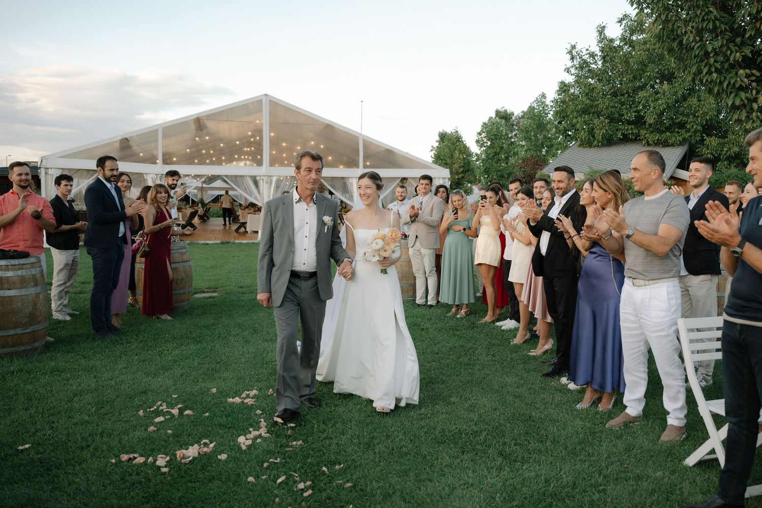Wedding guests gather outdoors under a tent during sunset for a celebration on a grassy lawn.