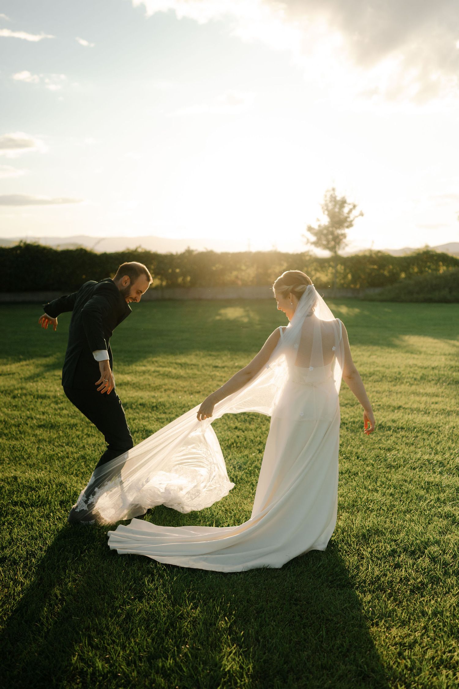 A couple in wedding attire plays with a flowing white dress train in a sunlit grassy field at sunset.
