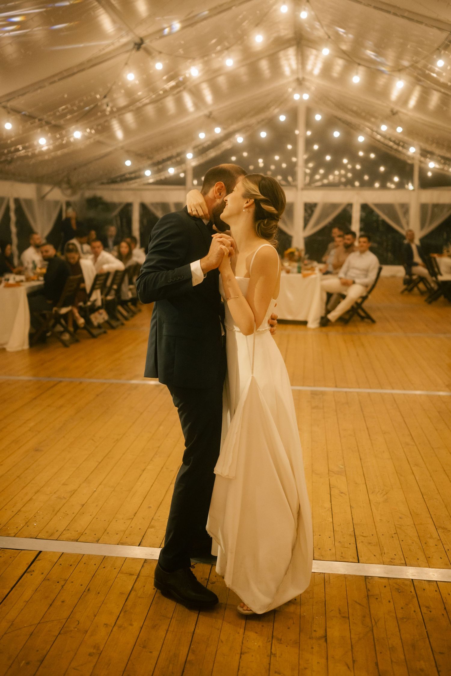 A couple shares their first dance at a wedding reception under twinkling lights in an elegant tent venue.