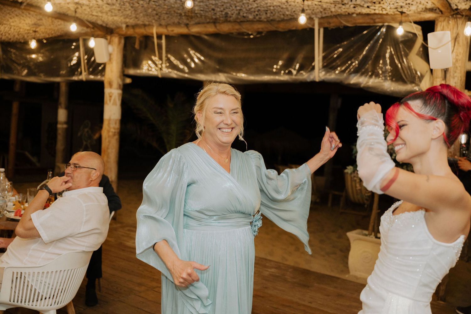 People dancing and celebrating at a rustic indoor reception venue with string lights overhead.