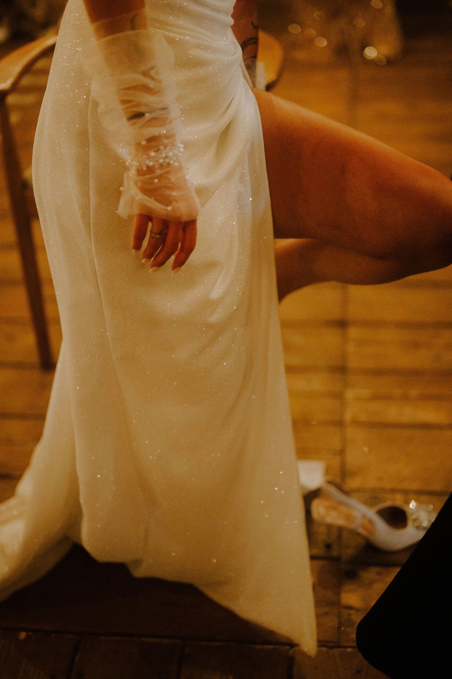 Close-up view of a bride's white wedding dress and hands against a warm-lit wooden floor.