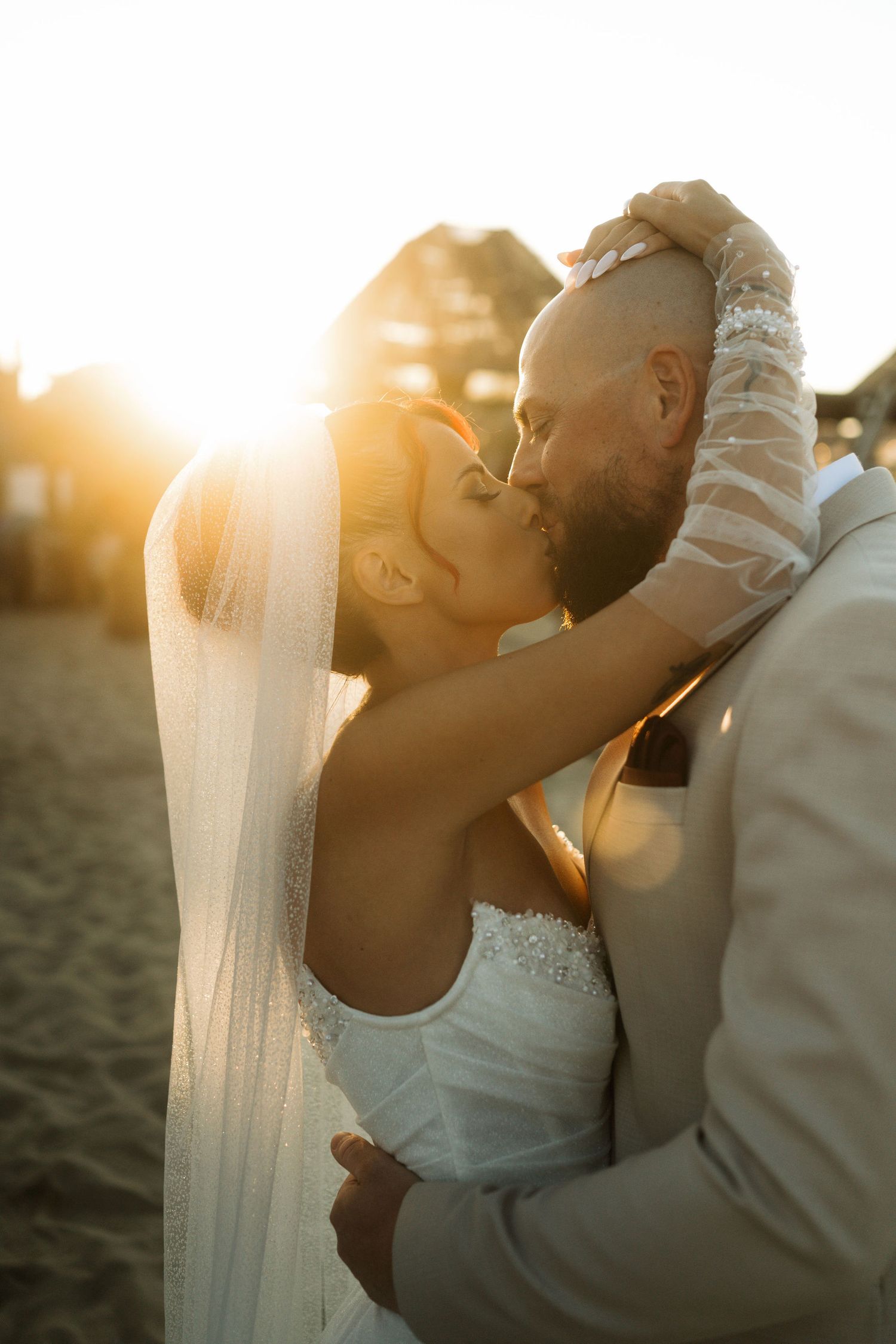 A couple shares a romantic kiss on the beach at sunset with golden light streaming through a wedding veil.