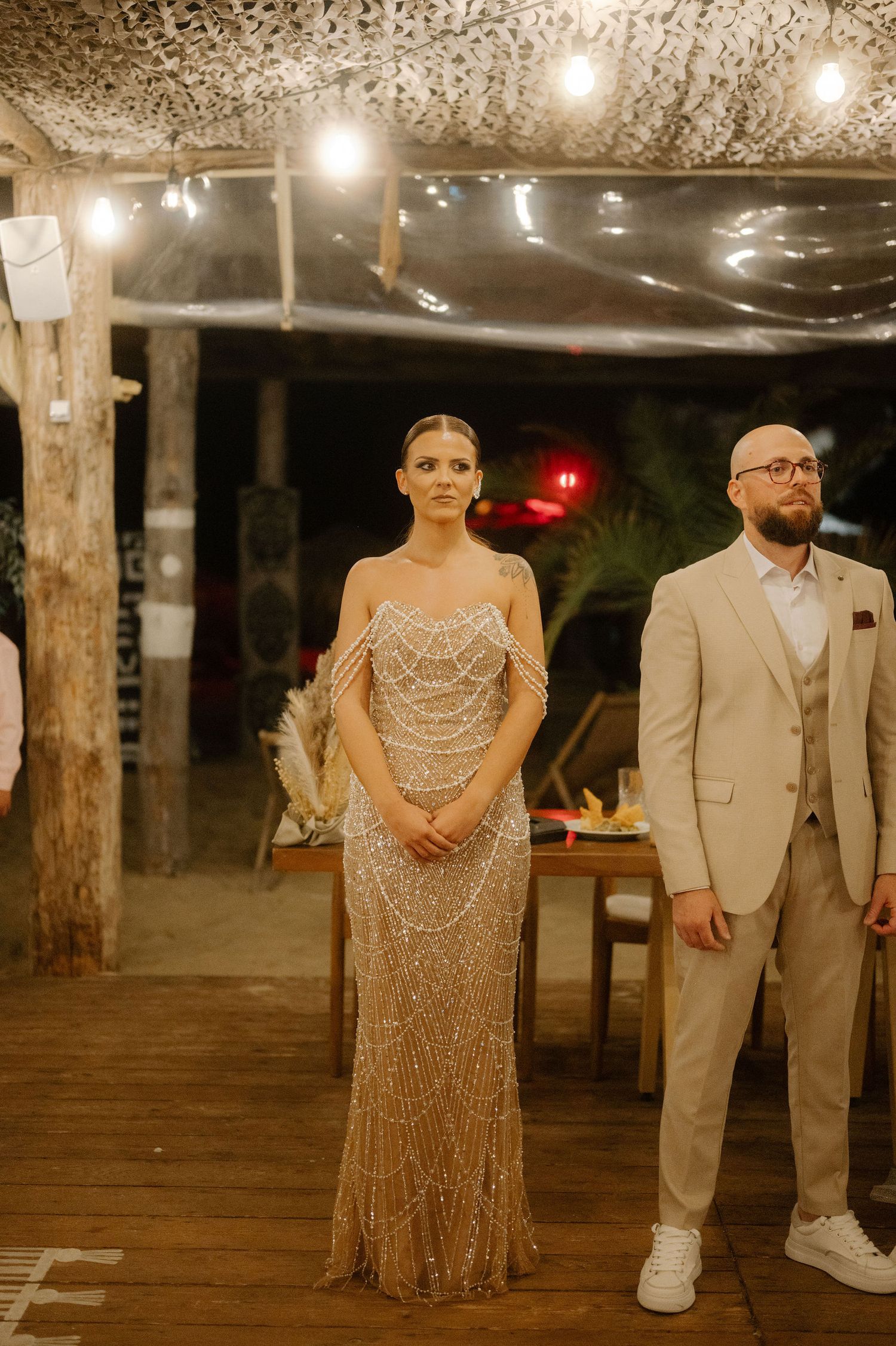 A couple stands at their wedding reception under string lights, with the bride in a sparkly gown and groom in a beige suit.