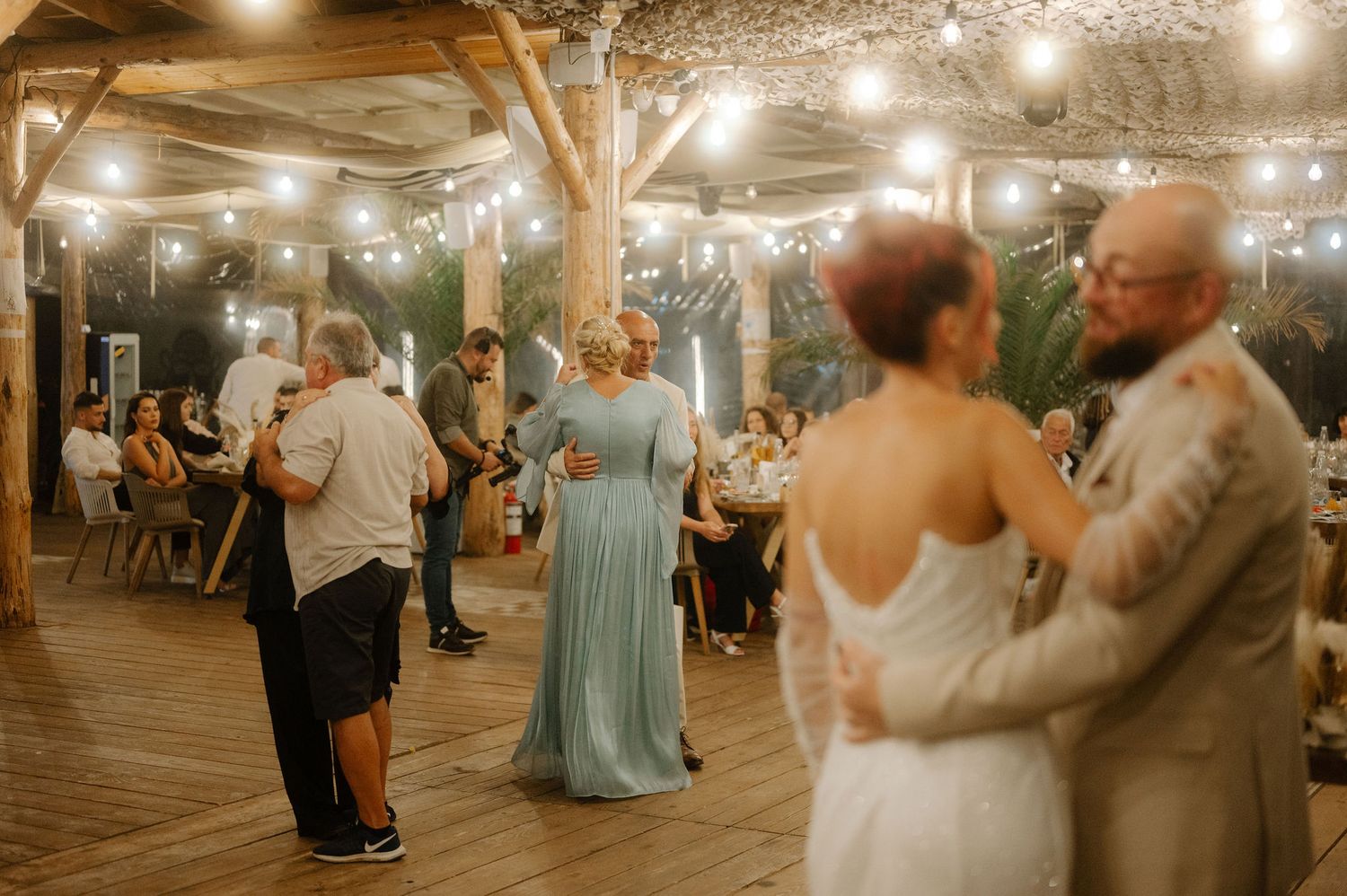 Couples dance together under warm string lights at a rustic outdoor reception venue with wooden beams overhead.