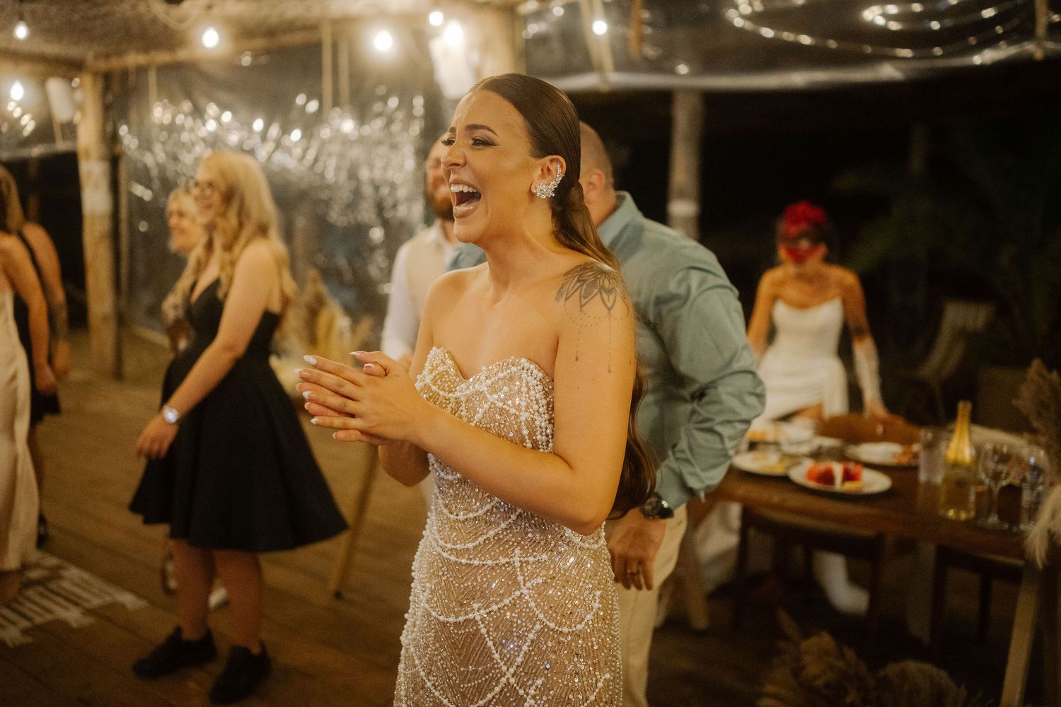 Bride laughs joyfully at outdoor evening wedding reception under string lights and dark sky.