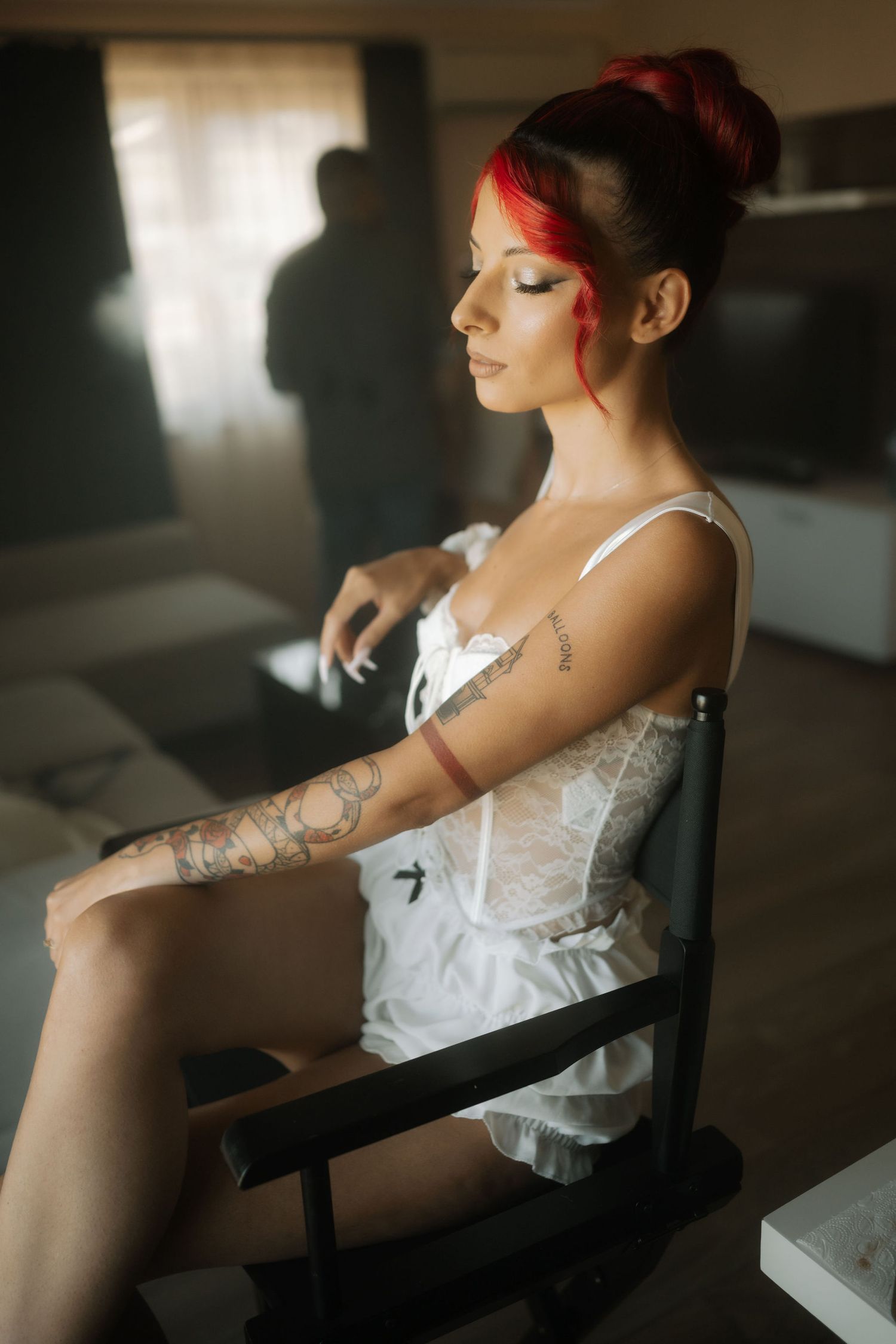 Artistic portrait in moody lighting shows a figure in white lingerie with tattoos and red hair styled in an updo.