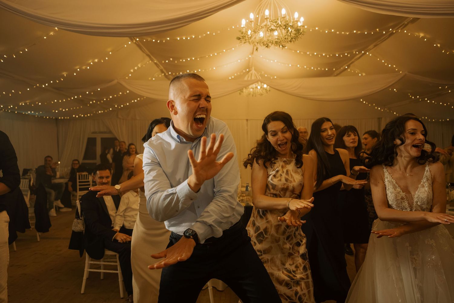 Groom dancing energetically at wedding reception with guests and bride laughing in background.