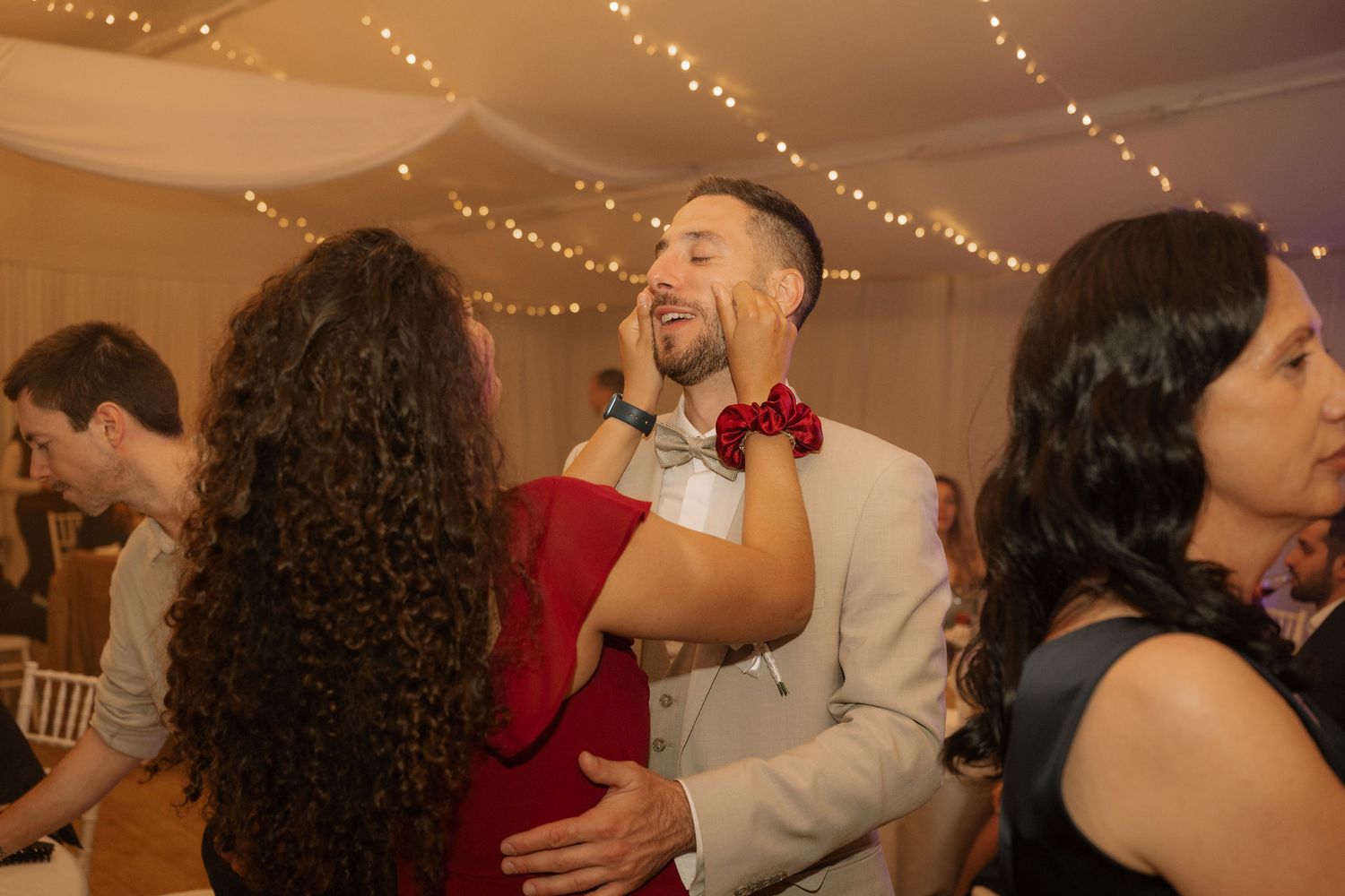 Couple dancing and laughing at a wedding reception with fairy lights and guests in the background.