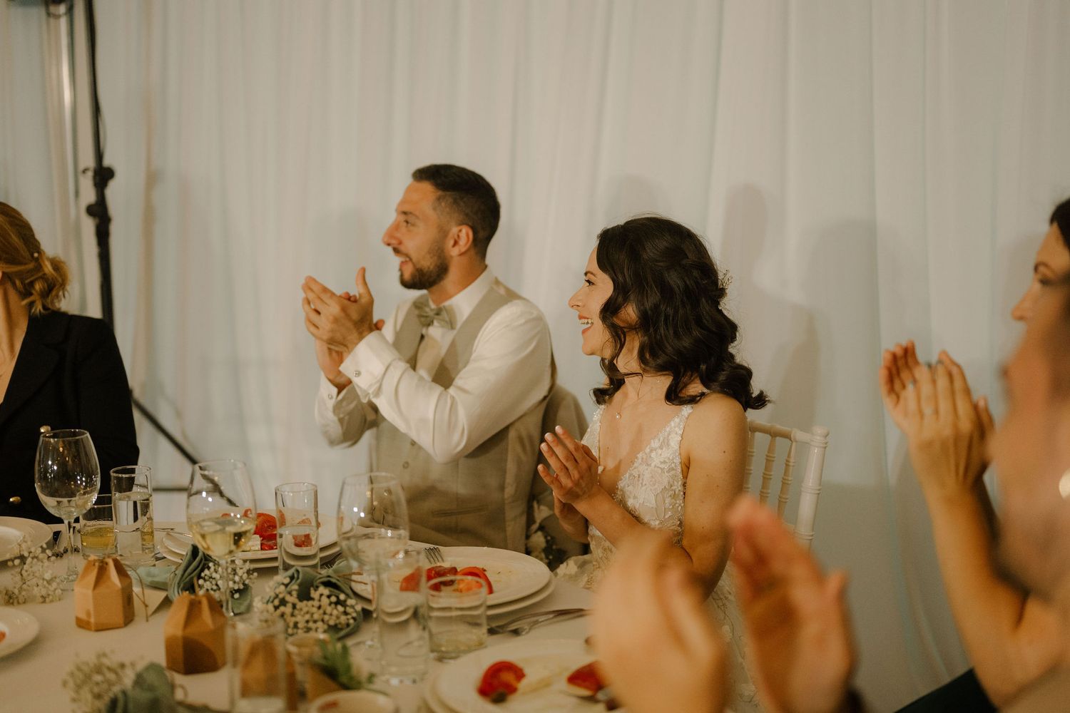 Bride and groom clapping at wedding reception table with guests, elegant white drape backdrop and floral centerpieces.