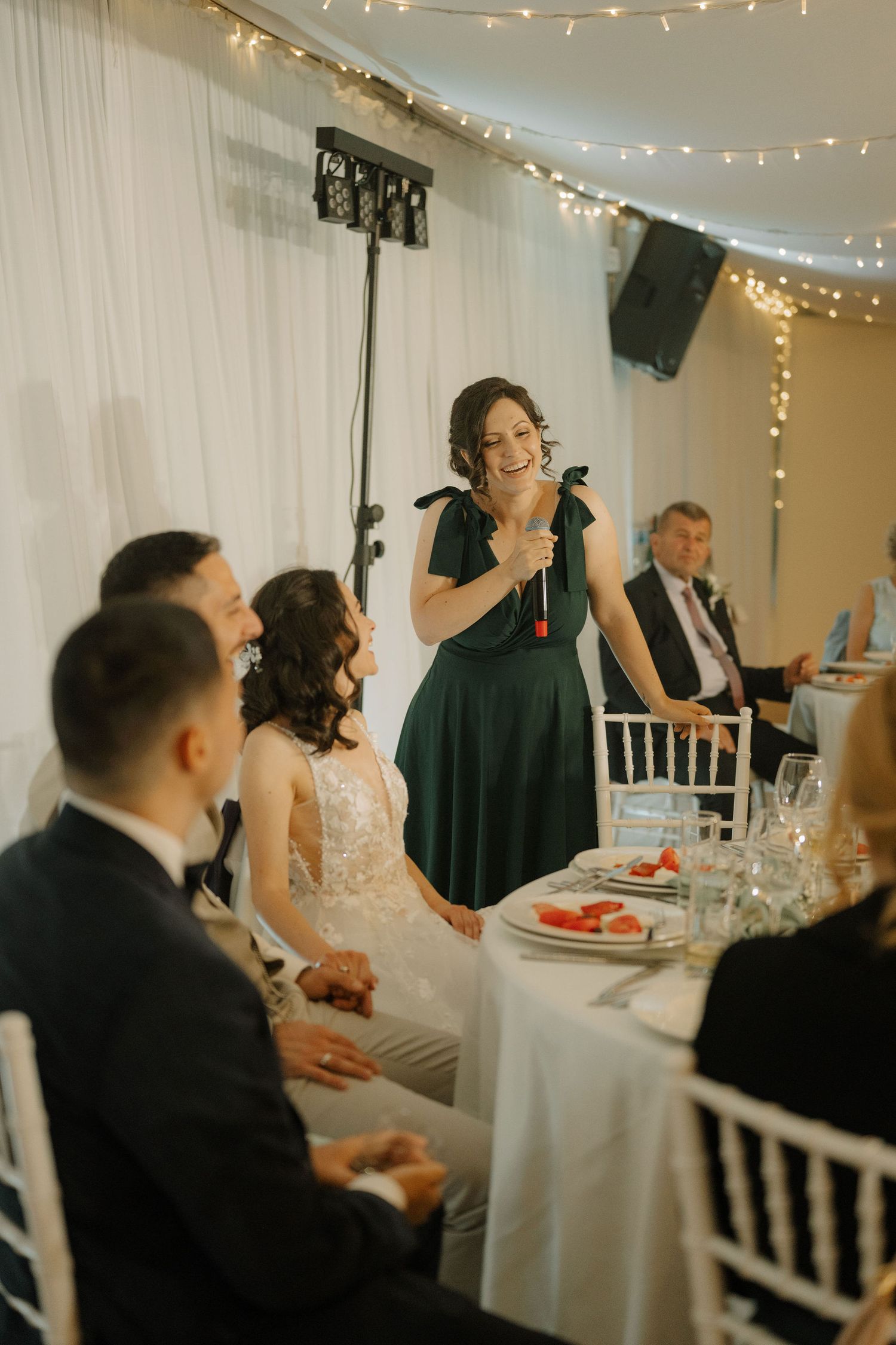A bridesmaid in a green dress gives a speech at a wedding reception while the bride and groom smile at a decorated table.