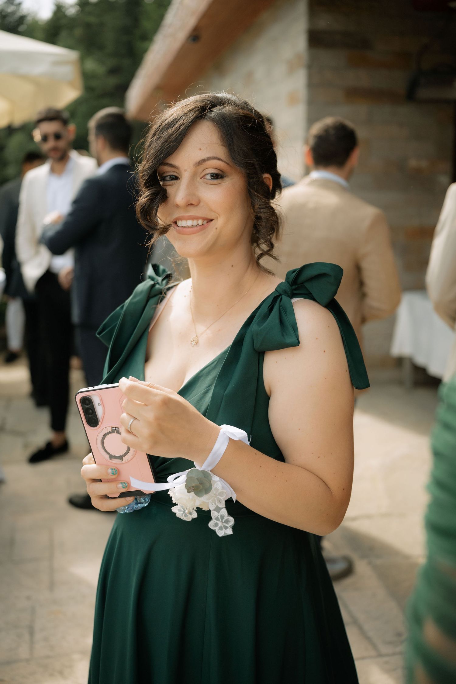 Woman in green dress holding pink phone smiles at outdoor wedding event.