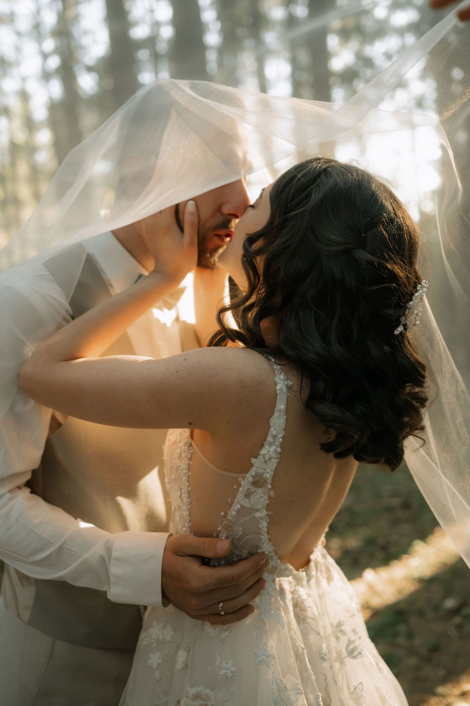Bride and groom sharing a romantic kiss under a flowing white veil in a sunlit forest wedding photo.