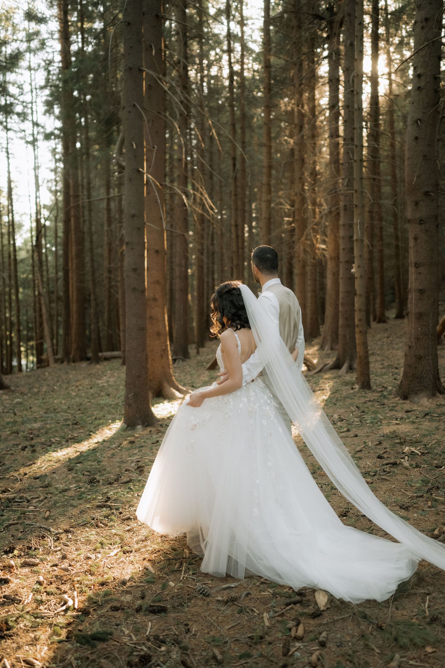 A bride and groom walk hand in hand through a sunlit forest, her white tulle gown and veil trailing behind them.