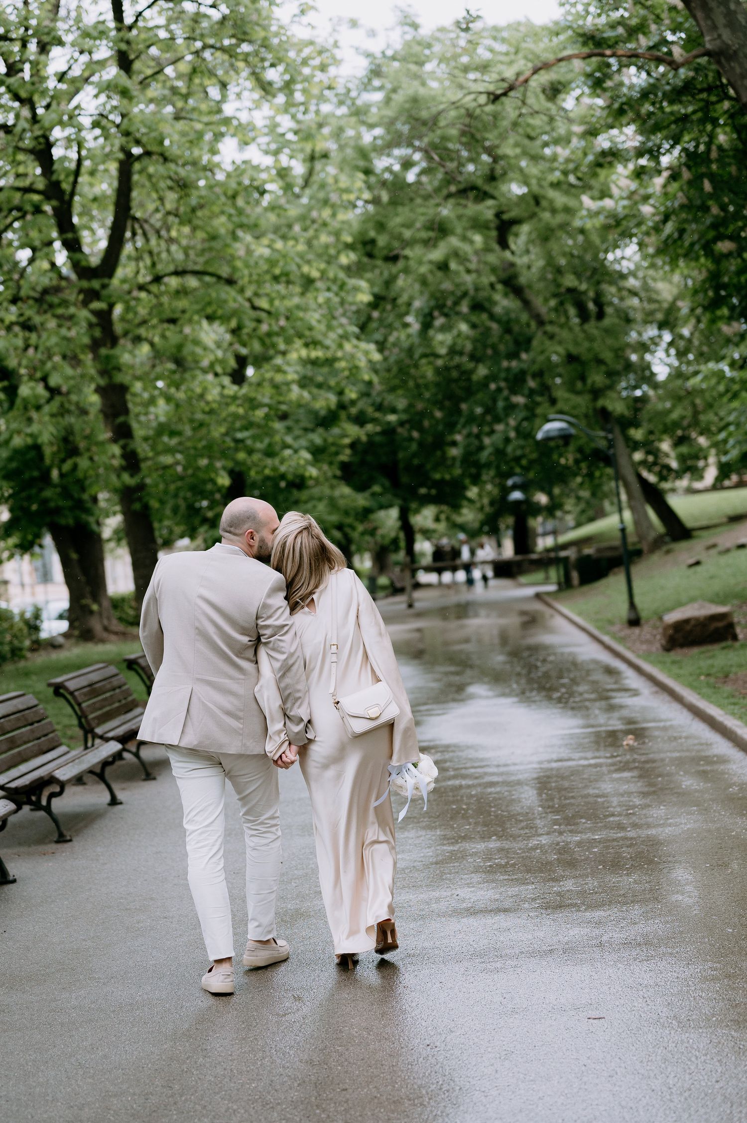 A couple walking arm in arm down a wet park pathway surrounded by green trees.