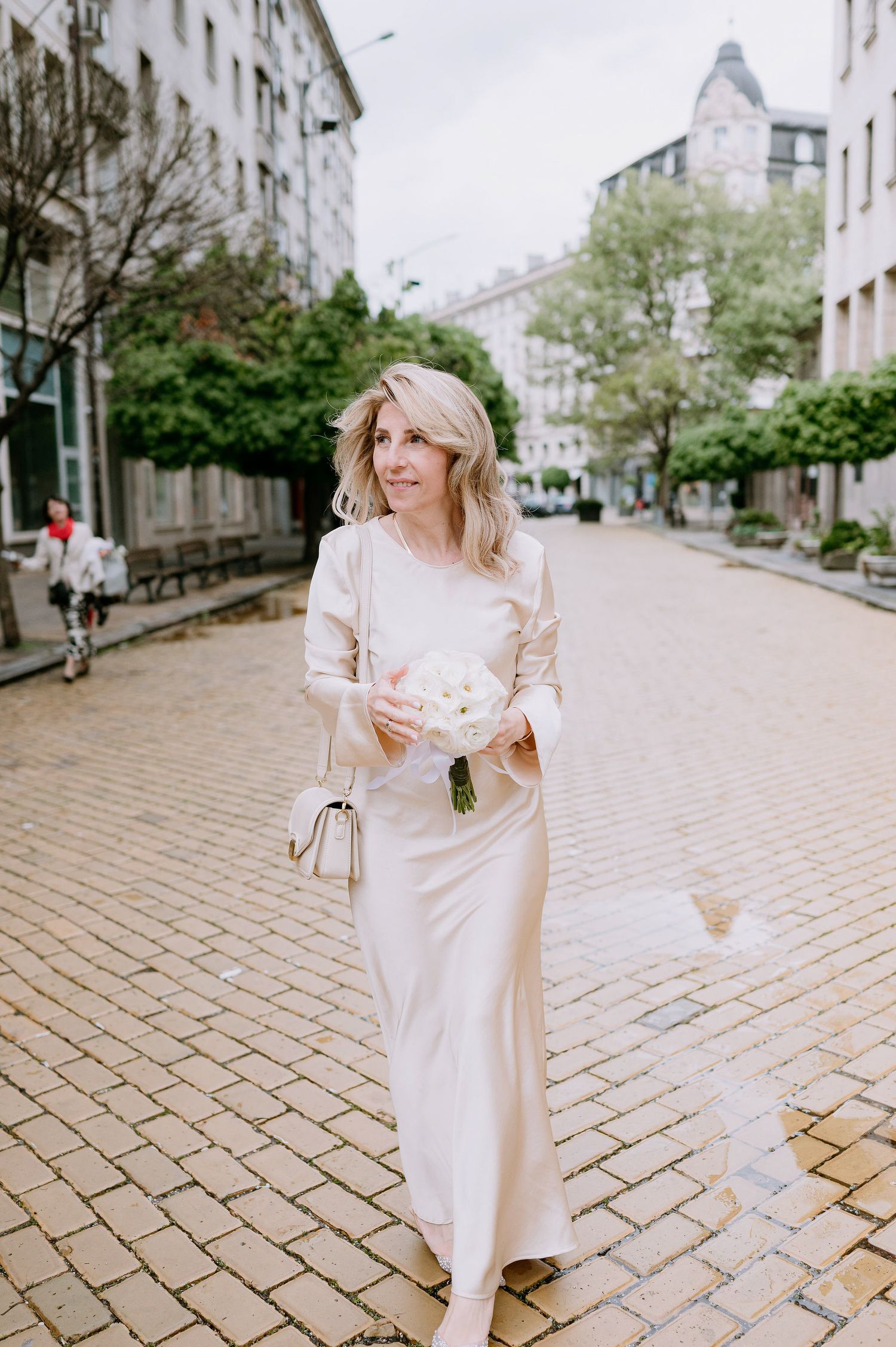 Person in flowing cream dress twirling on cobblestone street near historic buildings.