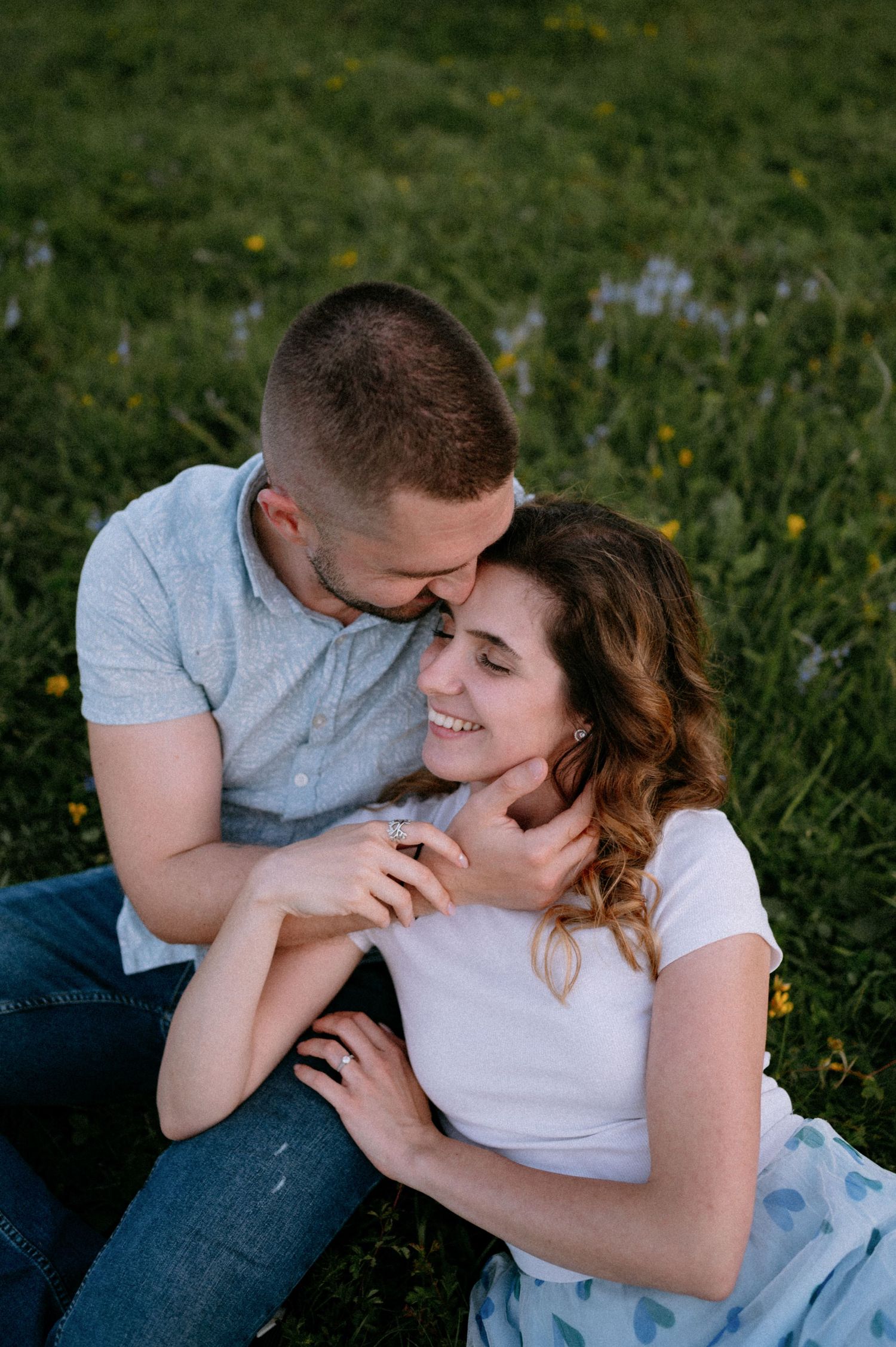 Joyful couple laughing together while cuddling on grass with yellow flowers.
