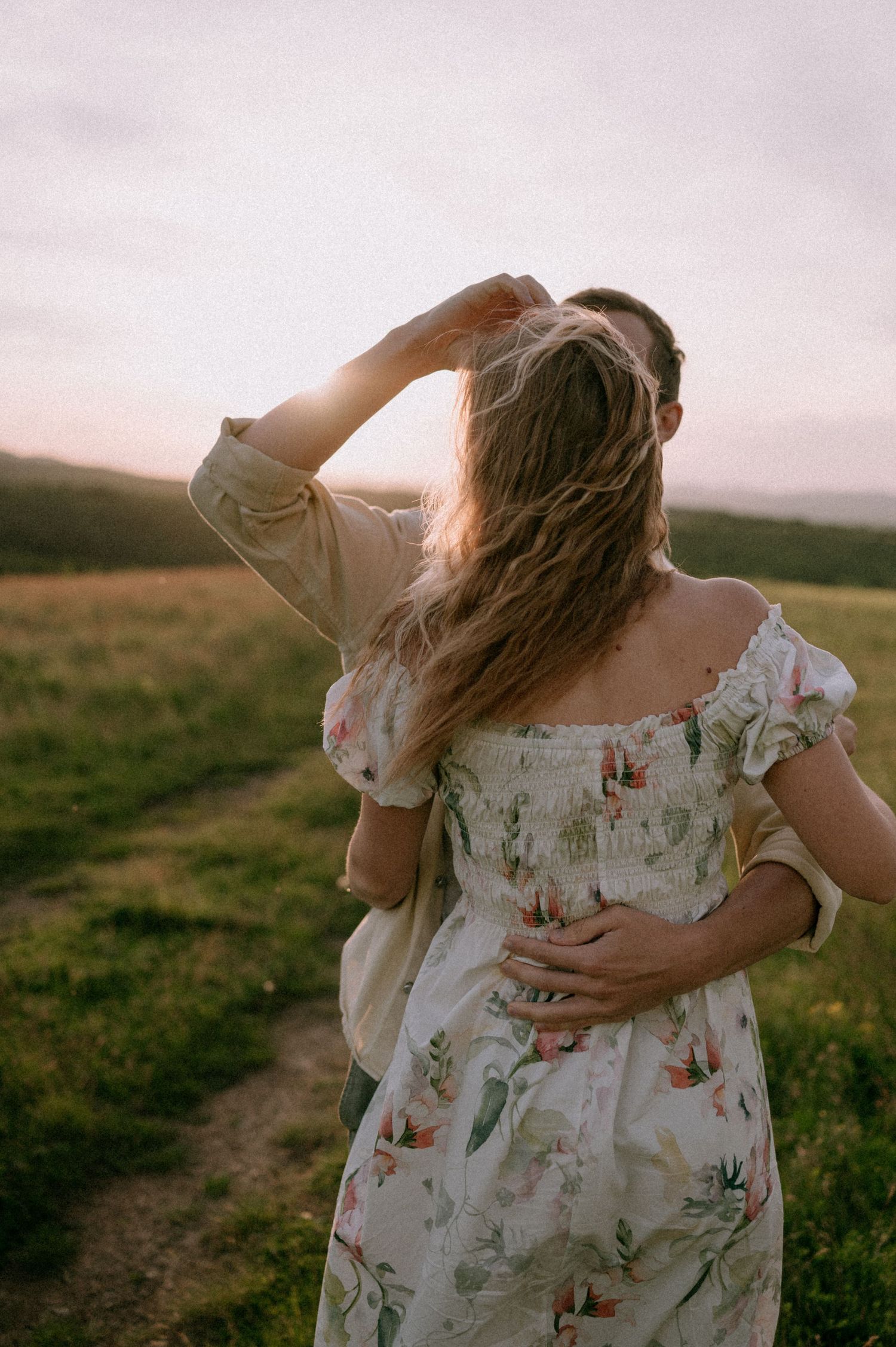 Two people share an intimate moment in a floral dress and cream shirt against a pastoral landscape.