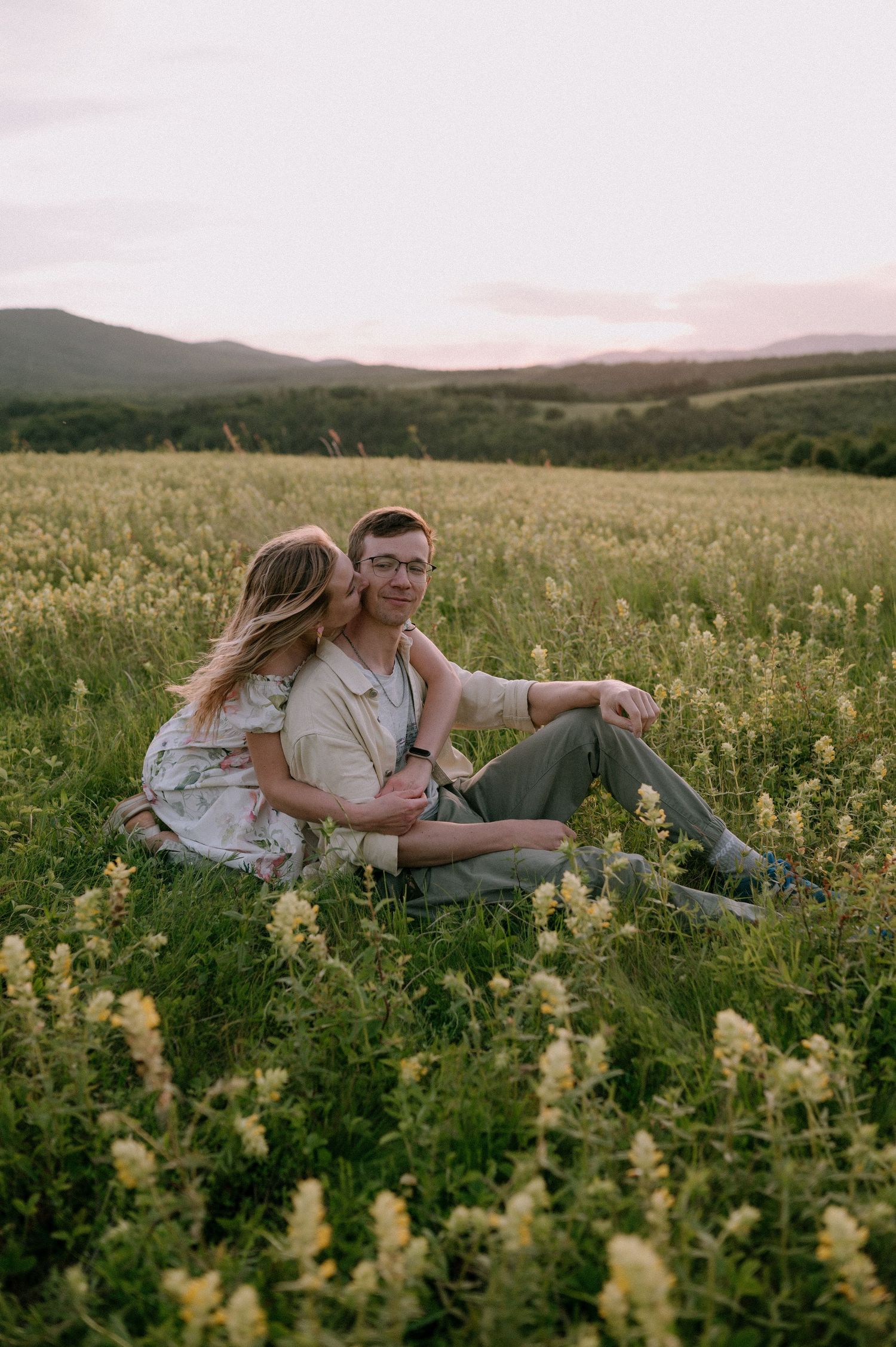 Couple relaxing together in scenic meadow with mountains and pink sky in background.