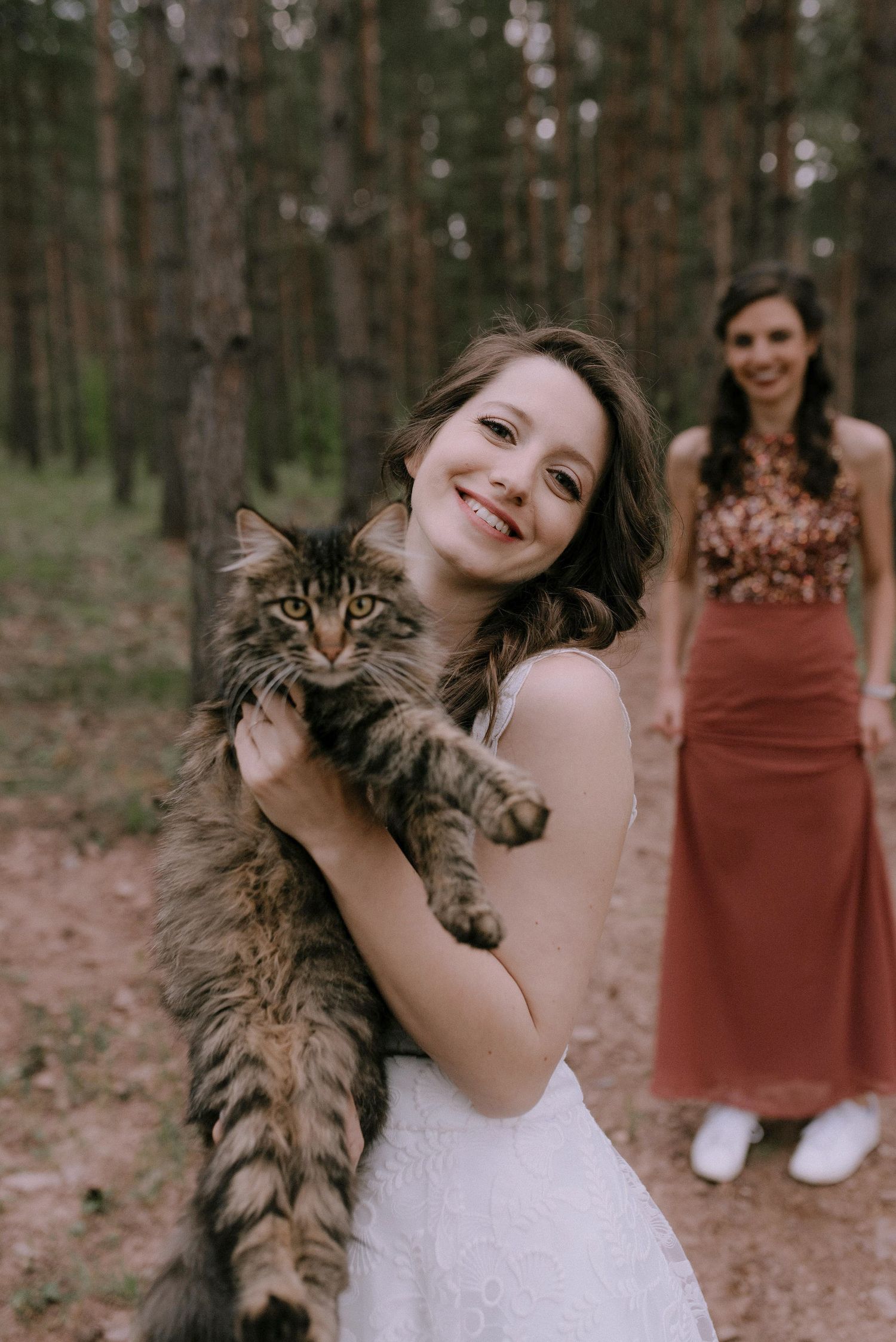 Person in white wedding dress holds and poses with a fluffy Maine Coon cat in the forest.