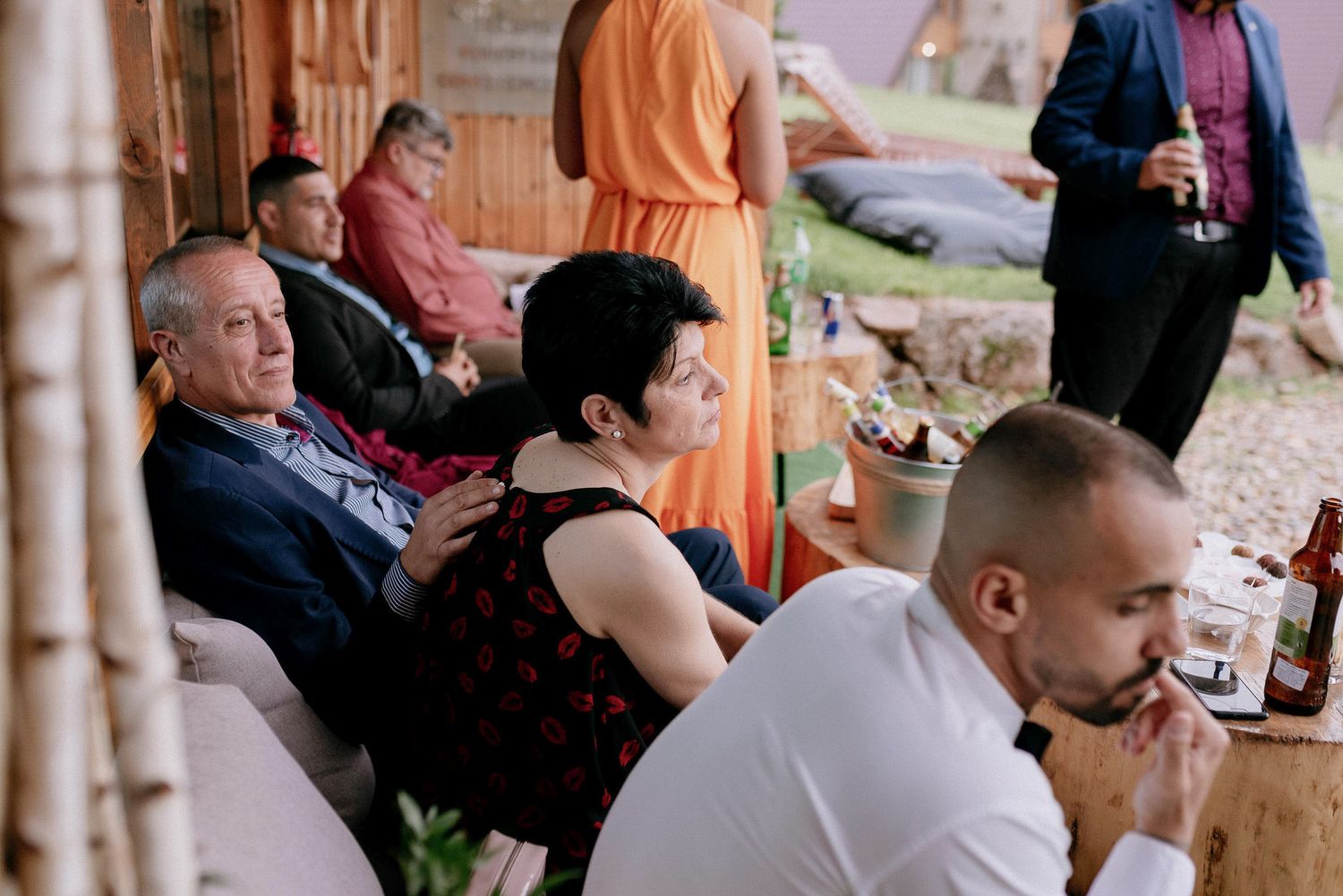 Wedding reception guests enjoying conversation at tables under wooden shelter.