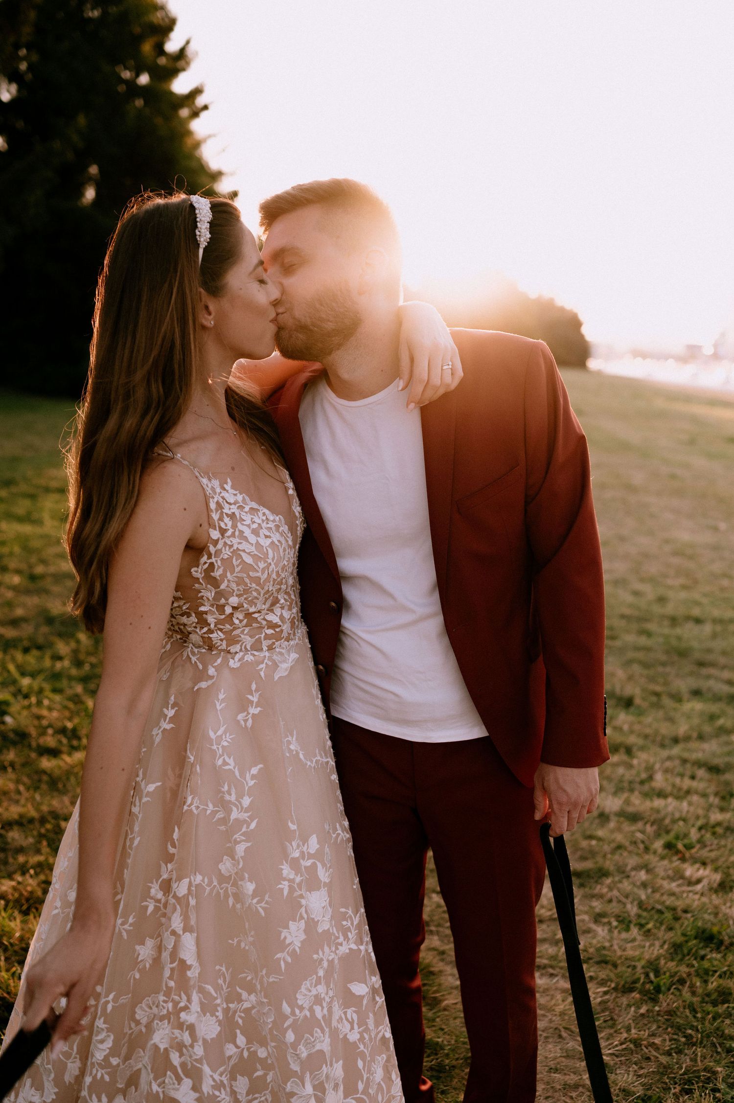A romantic sunset photoshoot with a couple in a white lace dress and burgundy suit embracing on a grassy hill.
