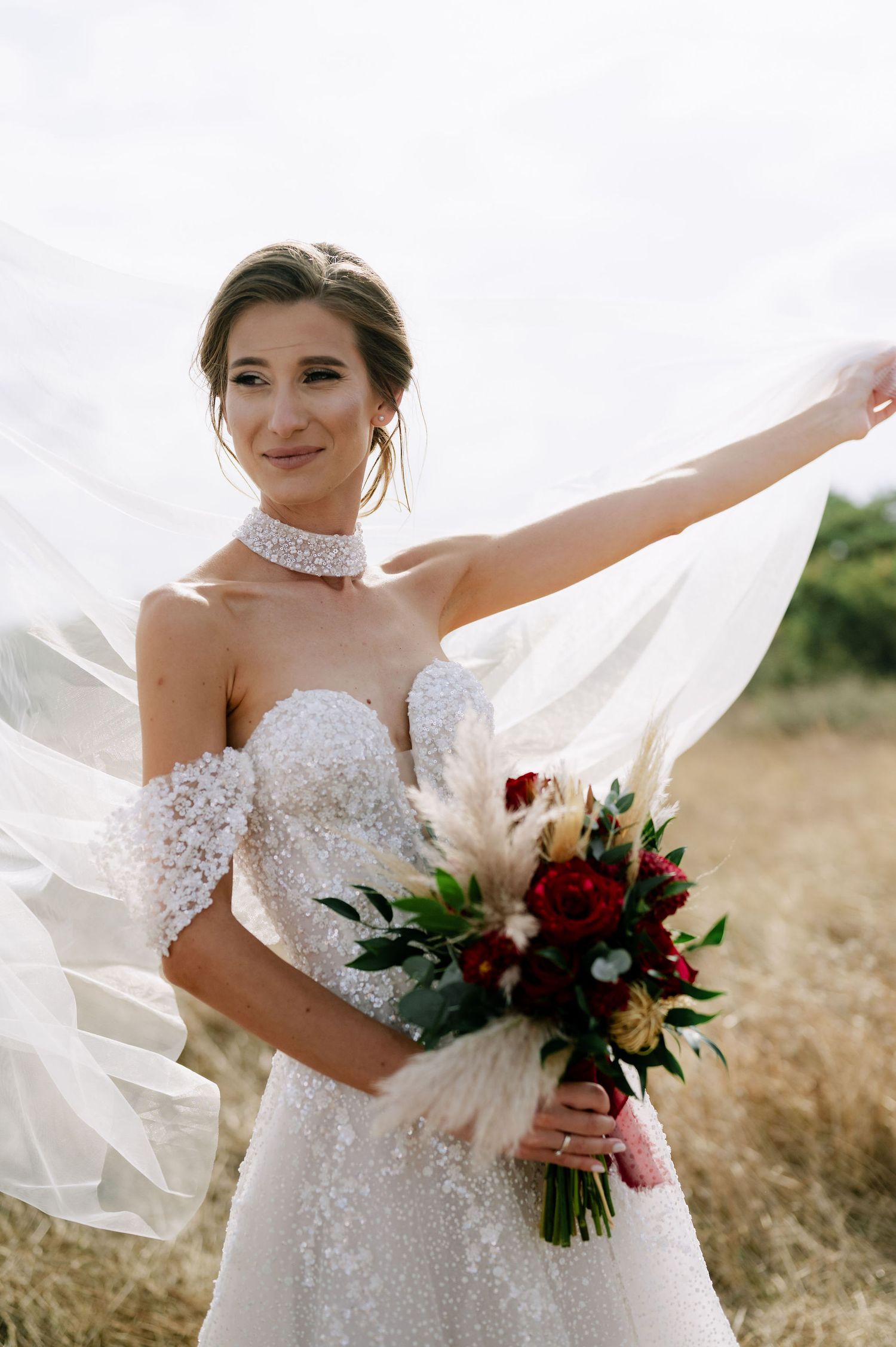 A bride wearing a beaded choker necklace holds her veil while carrying a burgundy and cream bouquet.