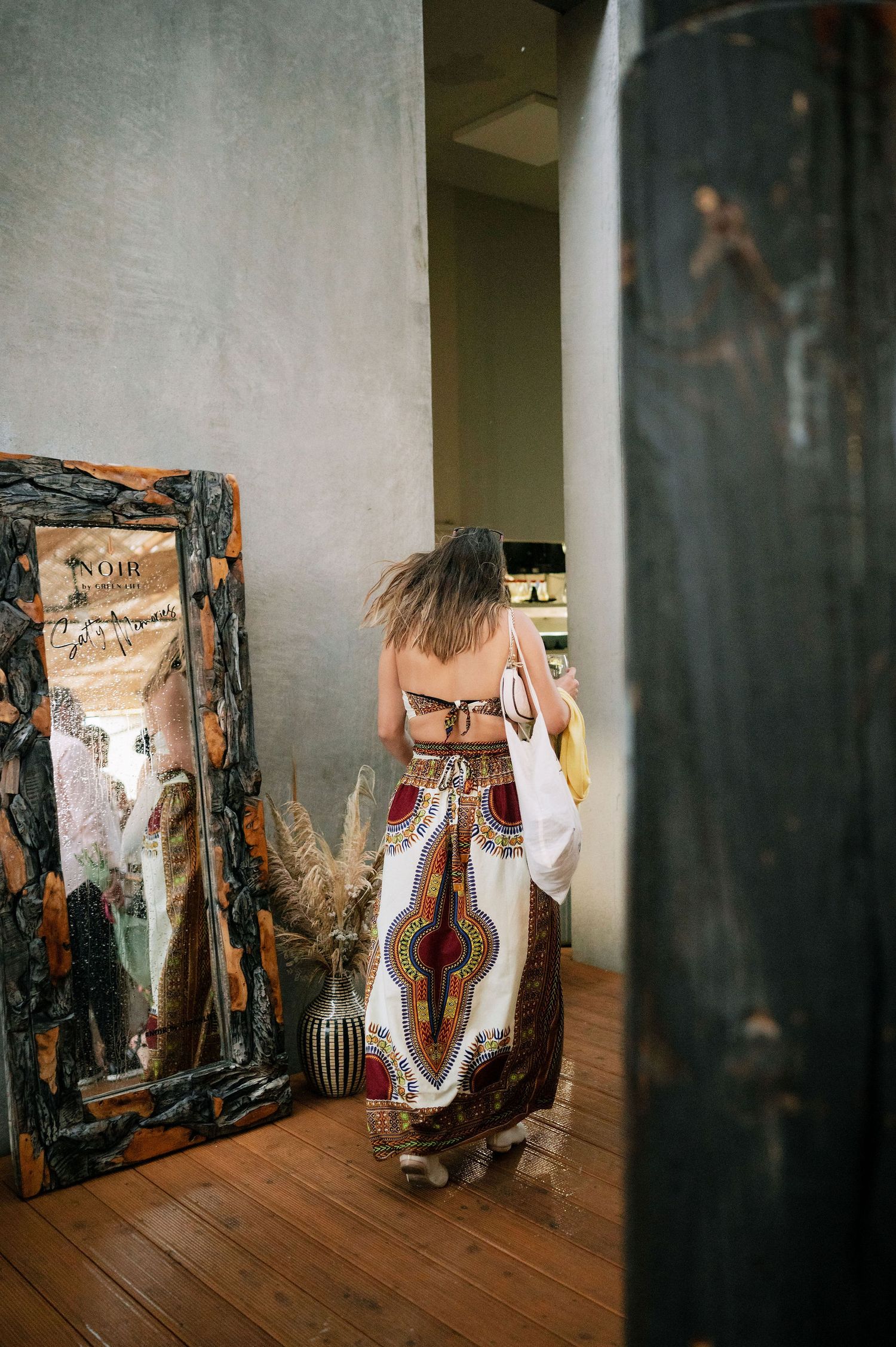 Person in patterned dress stands near vintage mirror in rustic indoor setting.