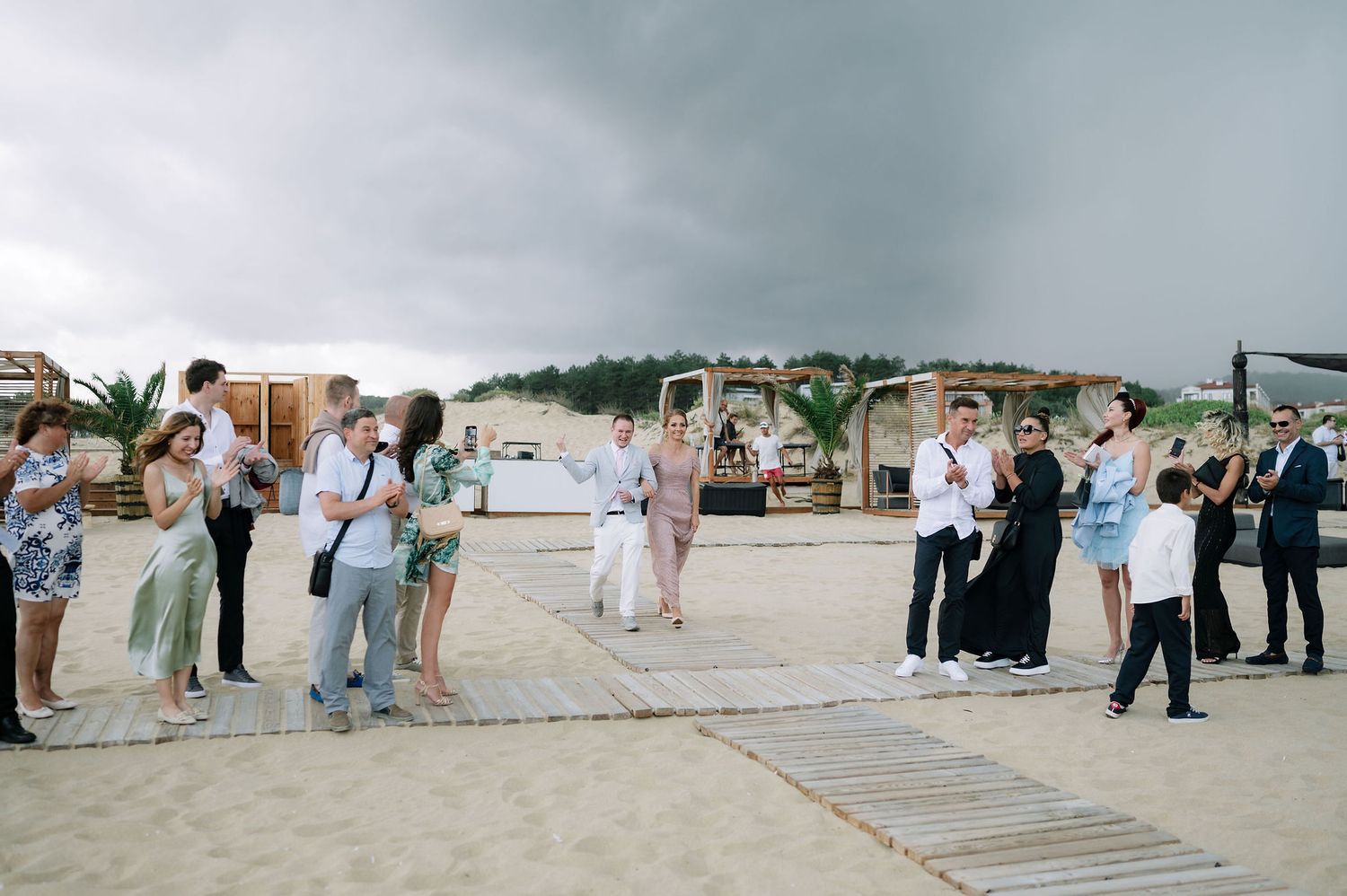 Wide angle shot of beach wedding guests lined up along a wooden boardwalk near the ocean.