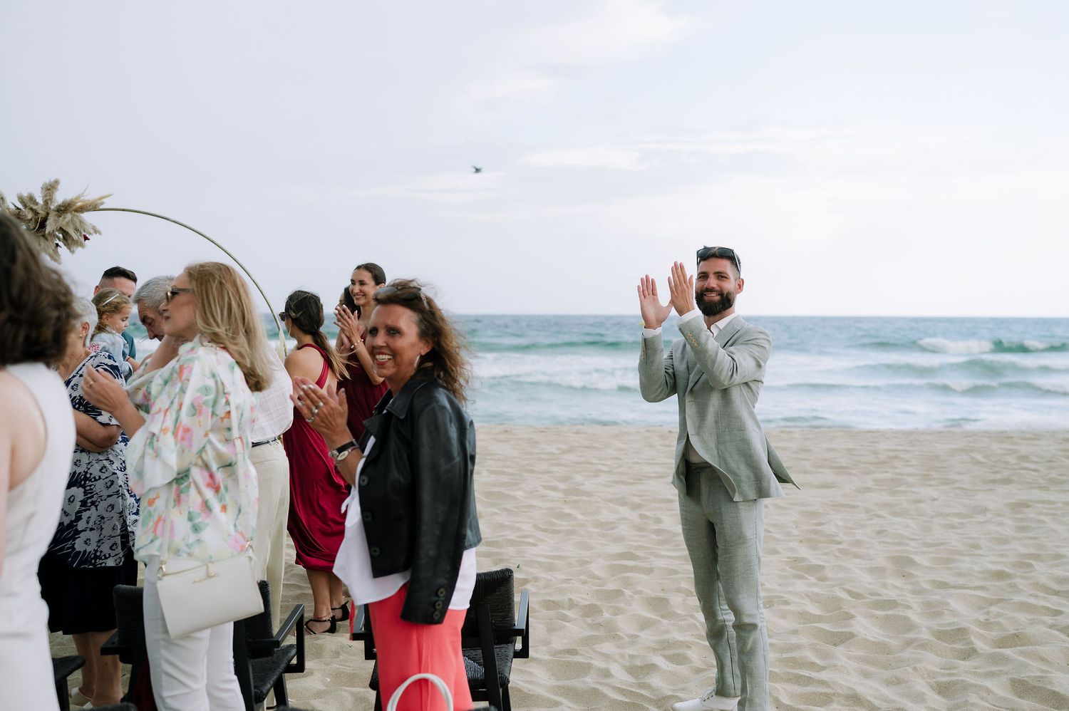 Wedding guests gather on a sandy beach boardwalk under cloudy skies for an outdoor celebration.