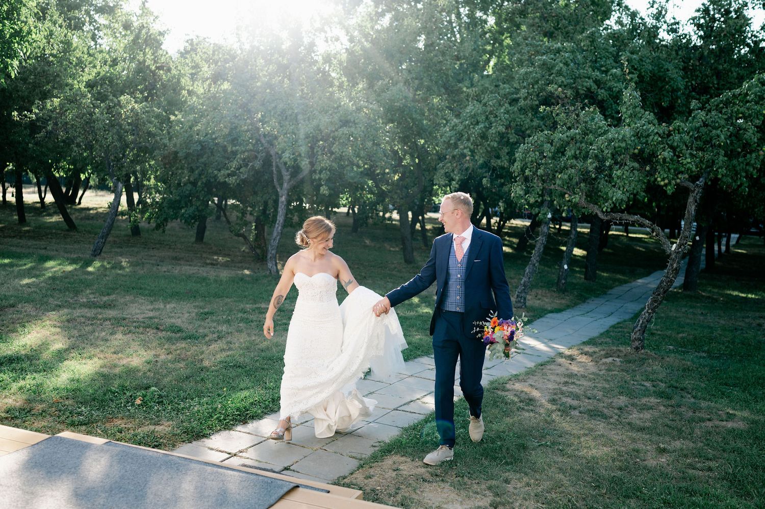 Newlyweds stroll down a scenic pathway with beautiful backlit trees and sunlight.