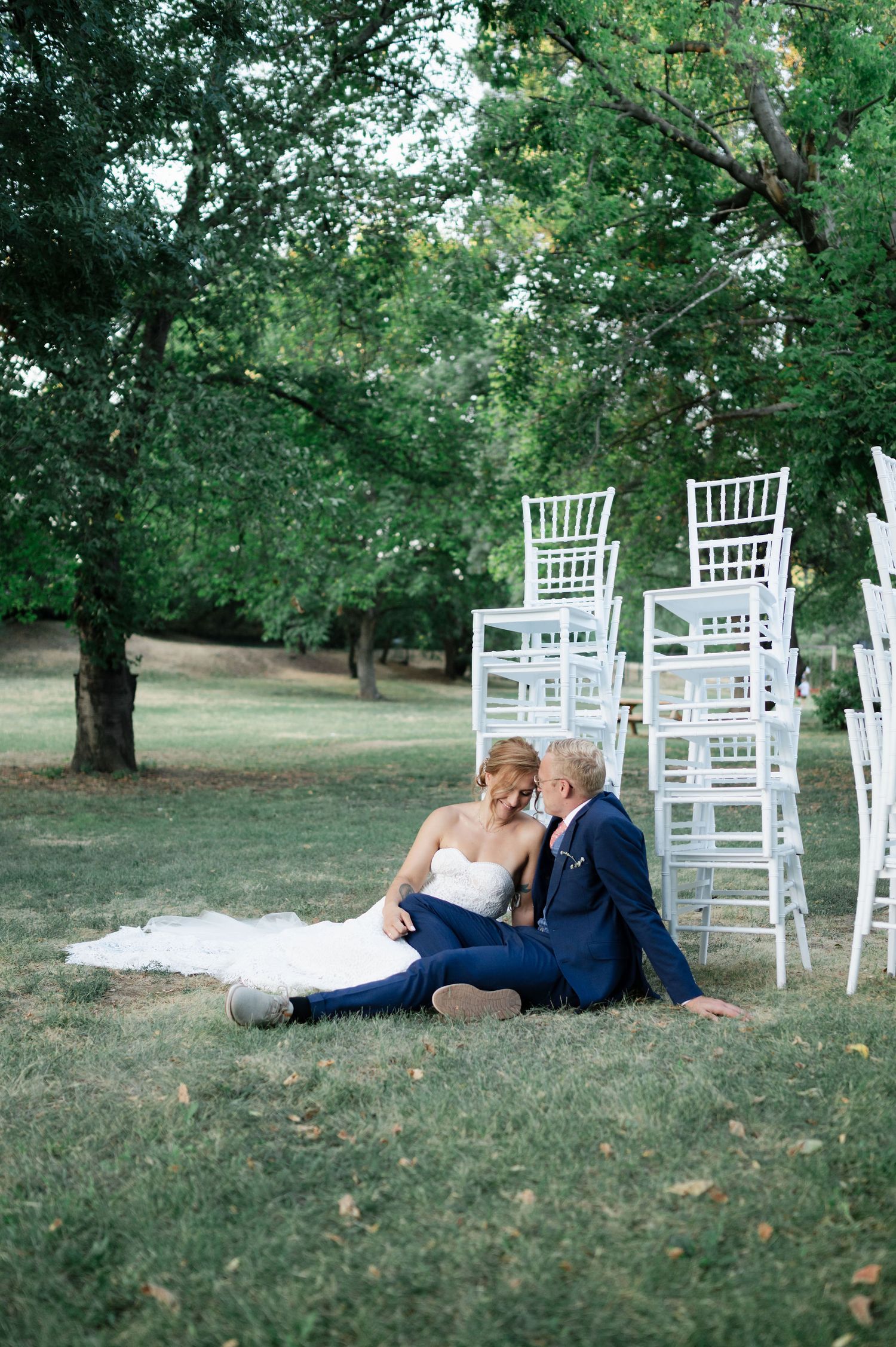 Newlyweds share tender moment on lawn with white ceremony chairs behind them.