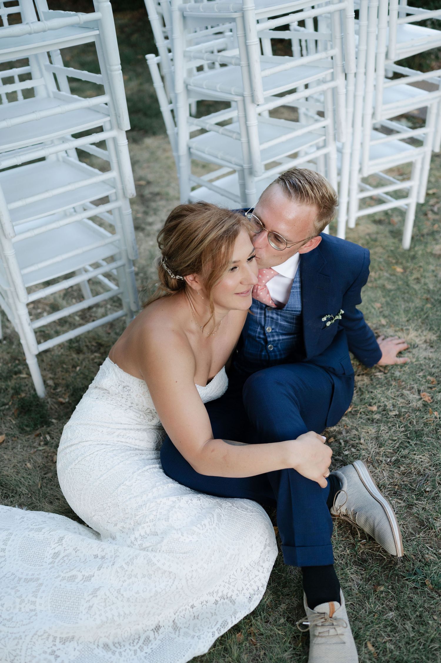 A couple in wedding attire shares an intimate moment on the grass near white chairs.