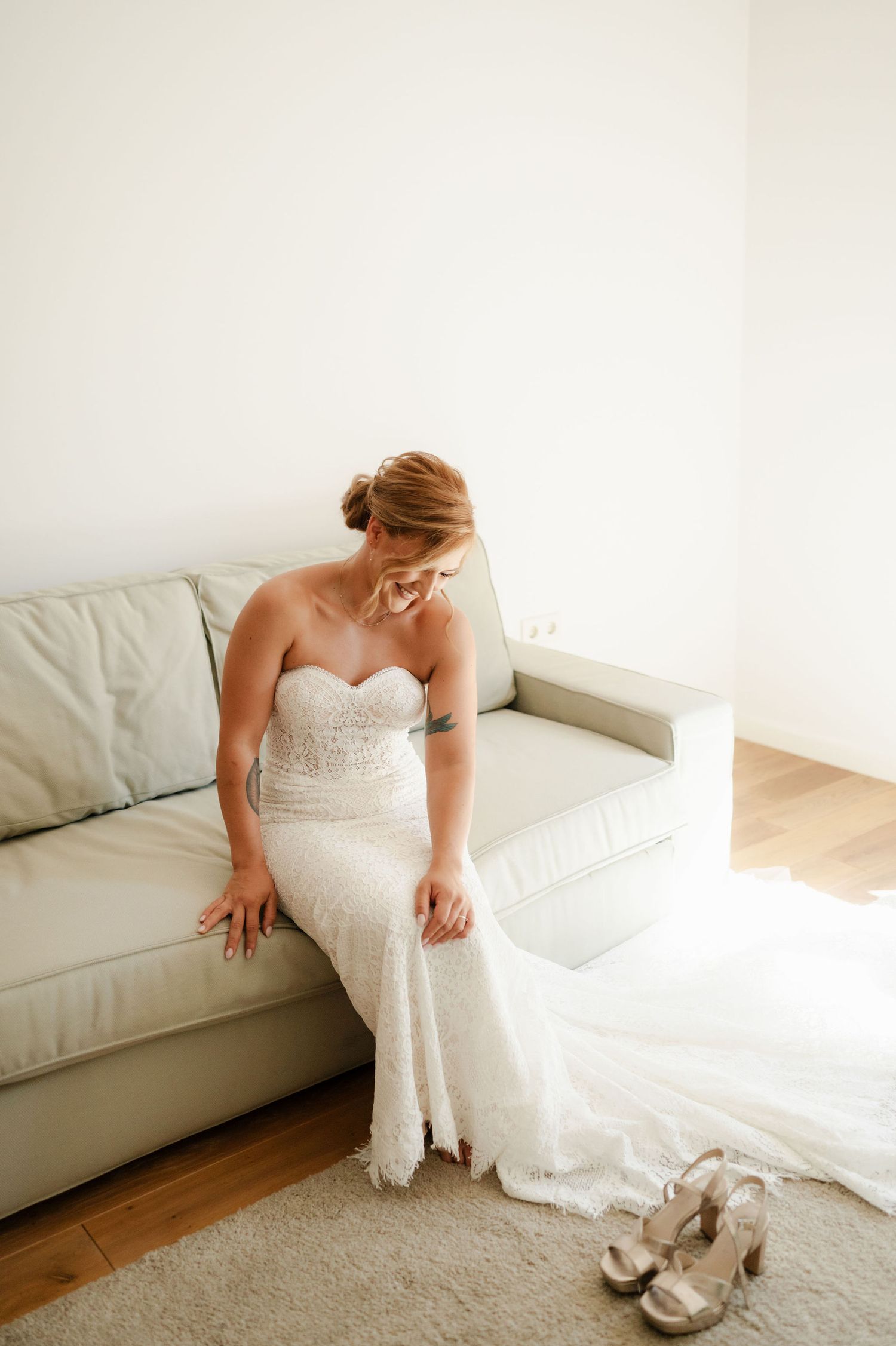 A bride sits on a white couch next to her wedding shoes in a bright room.