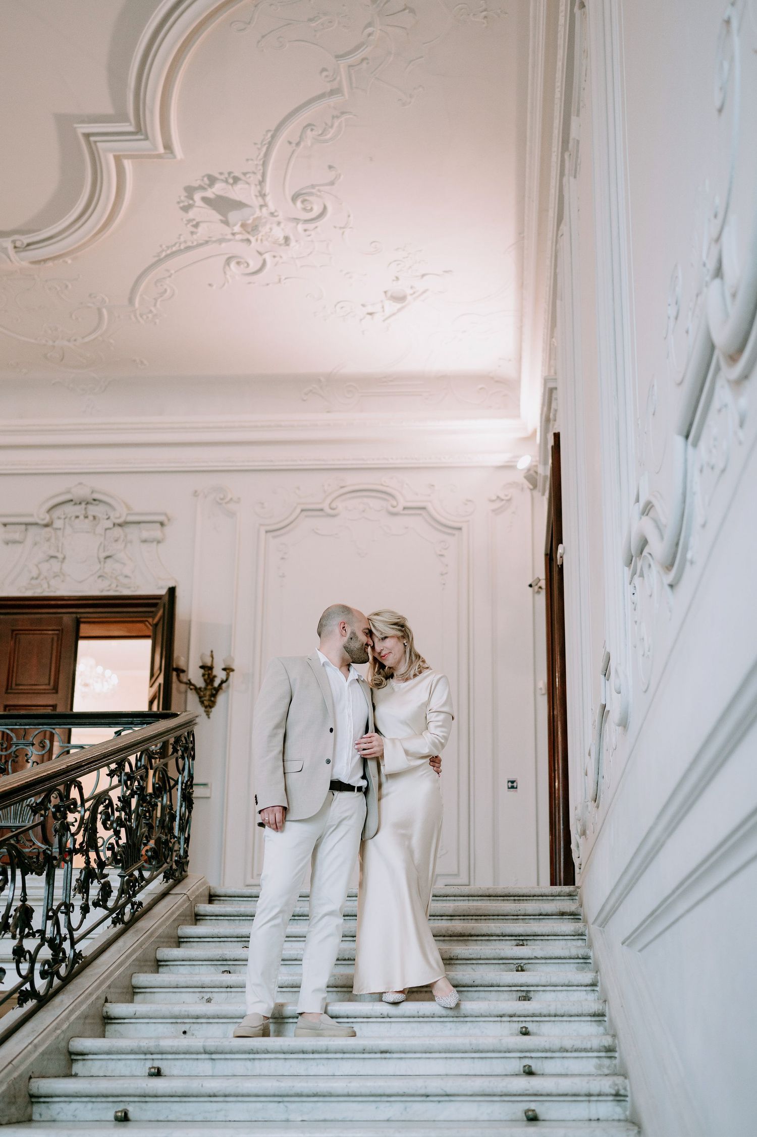 Elegant white staircase scene with ornate moldings and two people in white attire.