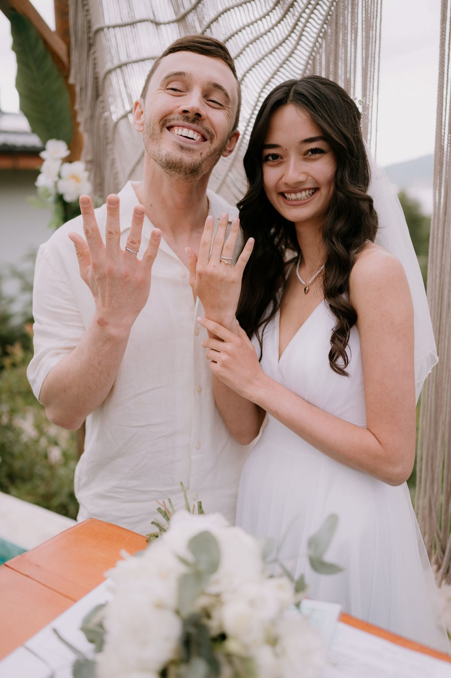 Couple in white outfits showing their rings while smiling at an outdoor venue.
