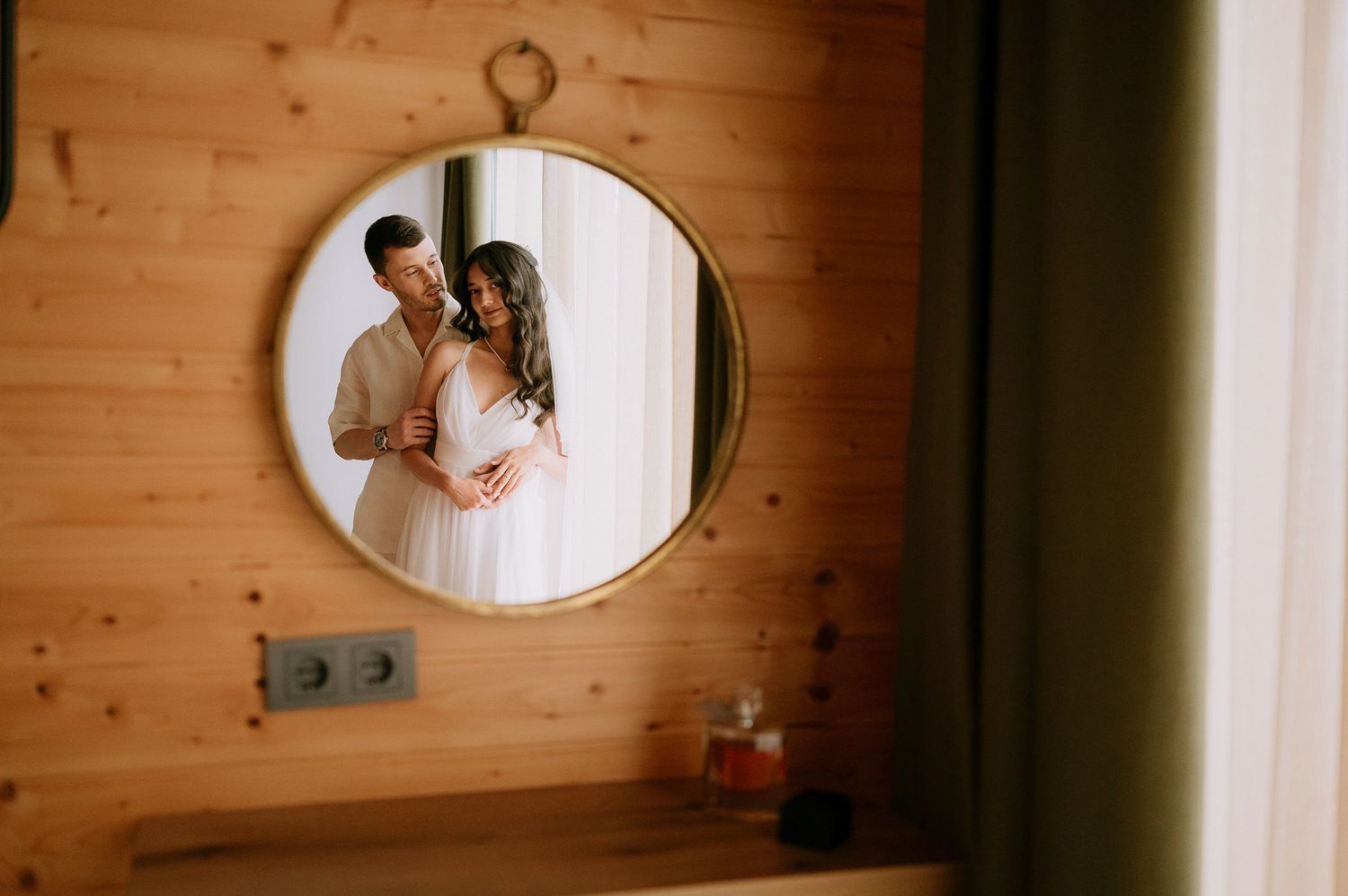 Wedding couple's reflection captured in a round mirror mounted on wooden cabin walls.