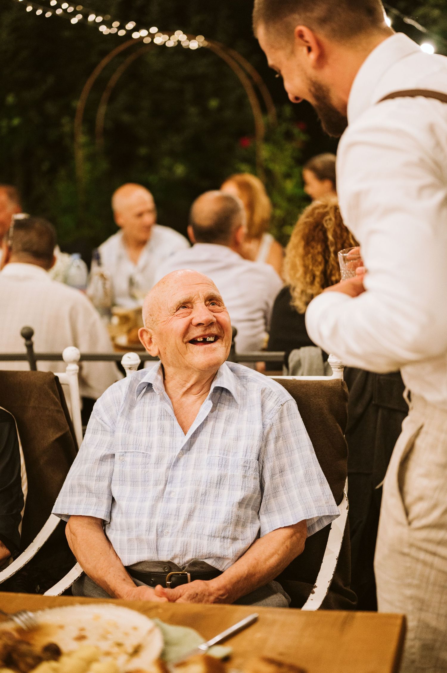 Elderly guest smiling warmly at an outdoor wedding reception under string lights.