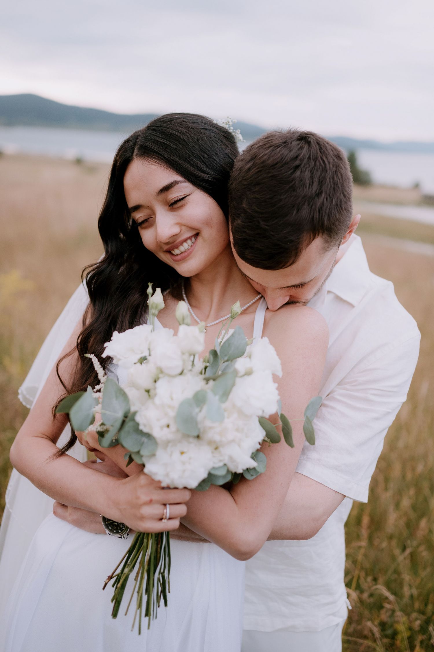 A romantic couple embraces in a field by the ocean while holding a white floral bouquet with eucalyptus.