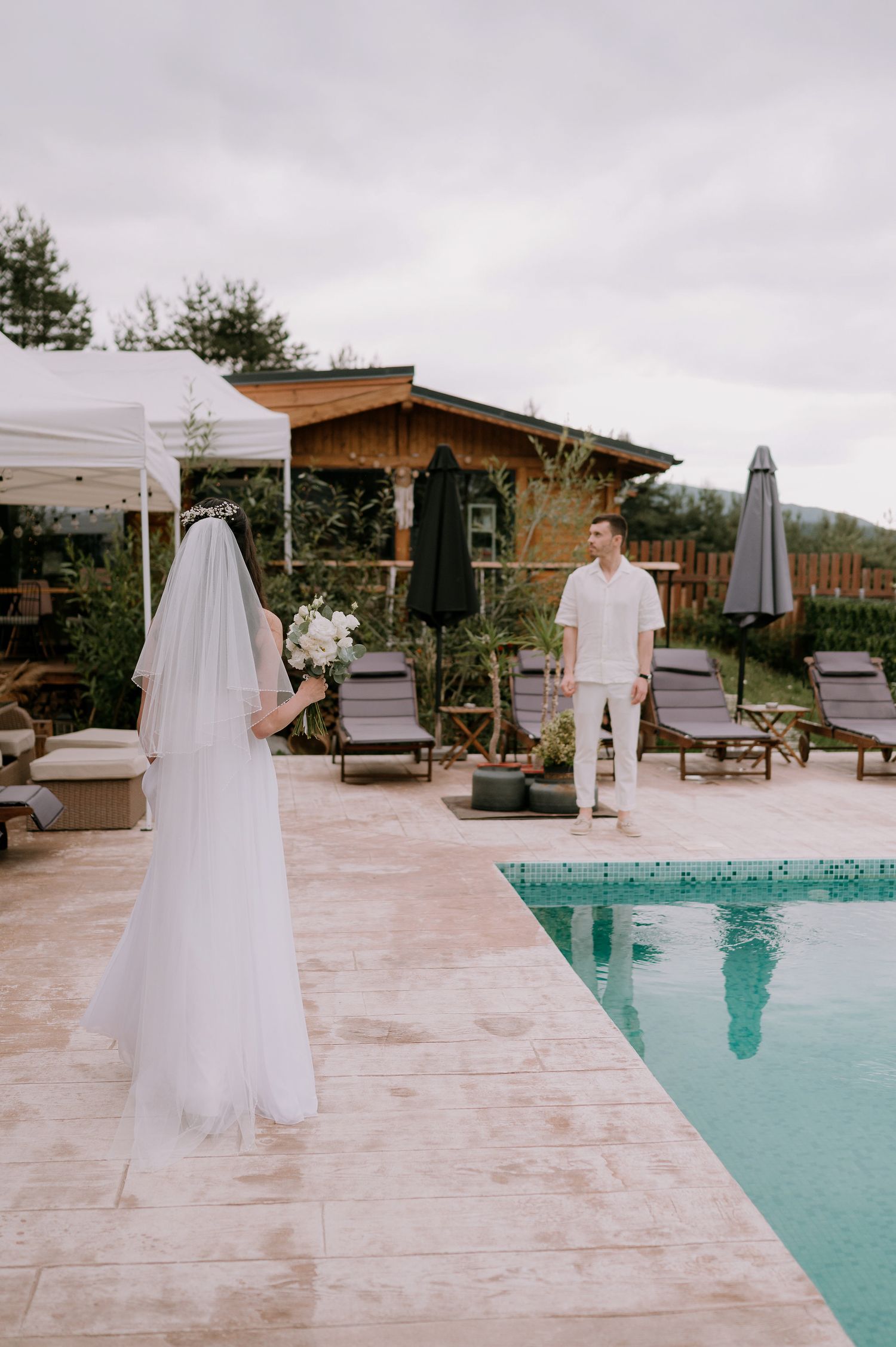 An elegant outdoor wedding scene unfolds beside a swimming pool with mountain views in the background.