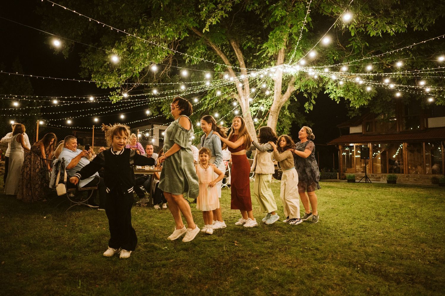 Wedding guests dance in a circle under twinkling string lights at an outdoor evening reception.
