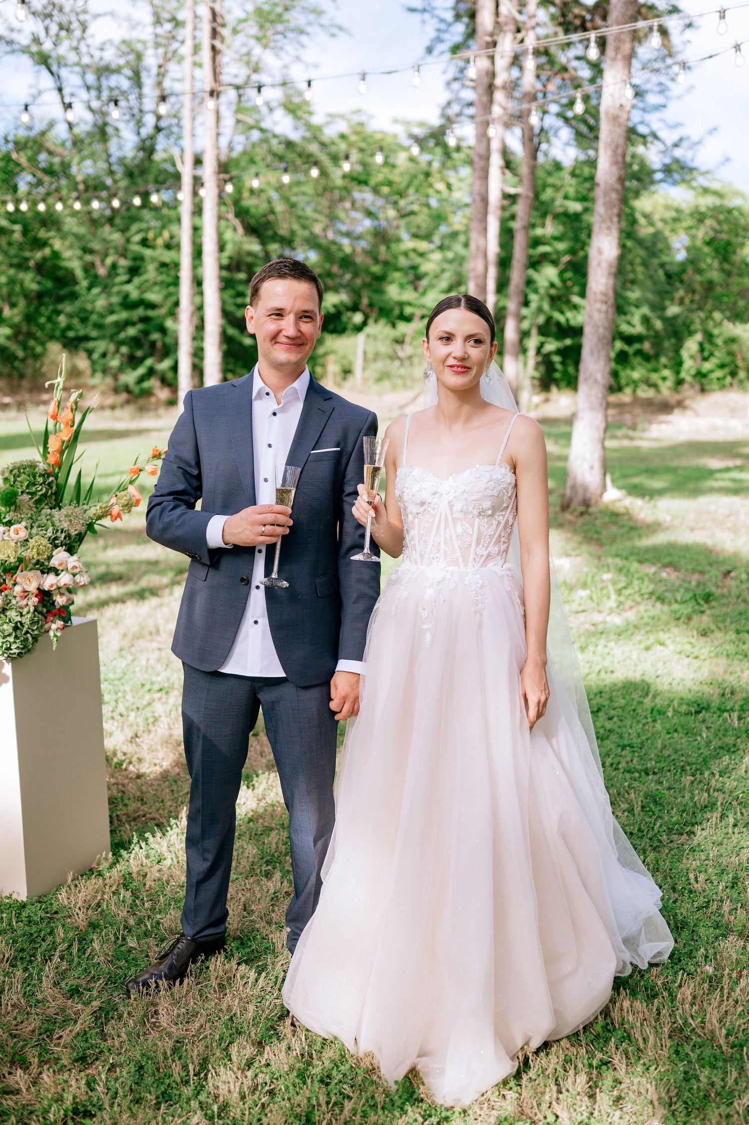 A couple shares a romantic moment in an outdoor wedding setting with tall trees and floral decorations.
