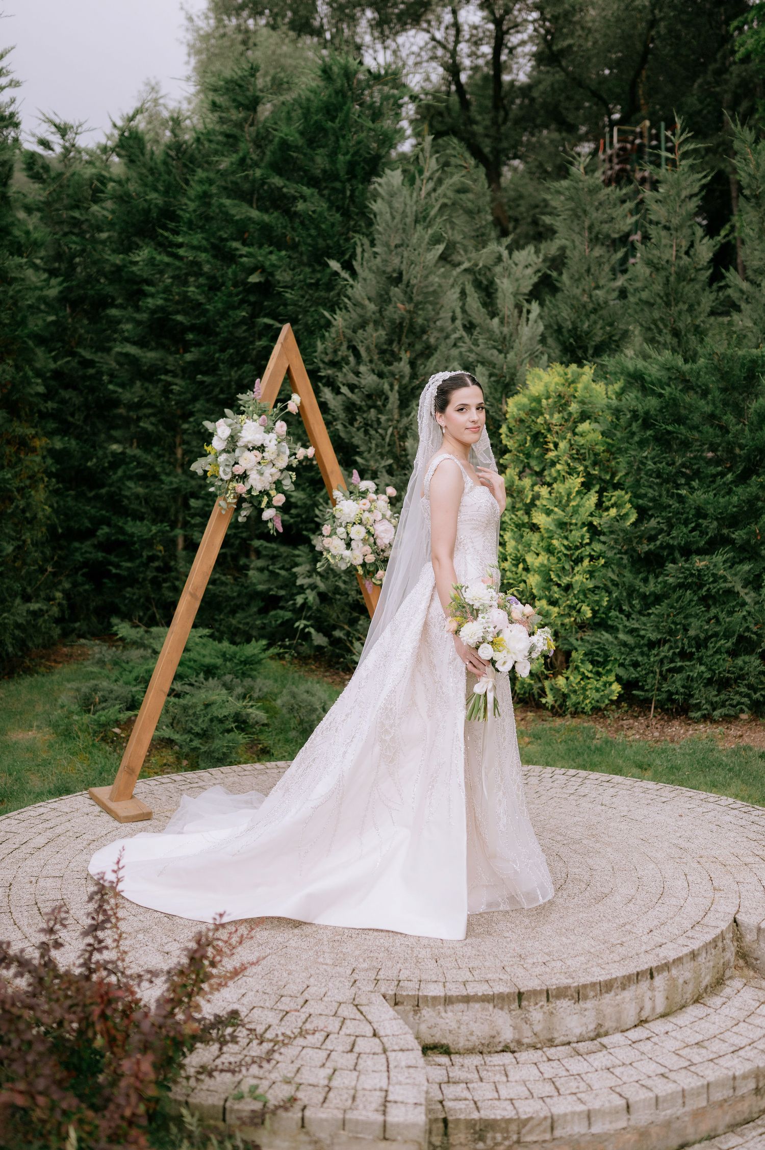 Bride in flowing white gown standing on circular stone platform with wooden ceremony arch.