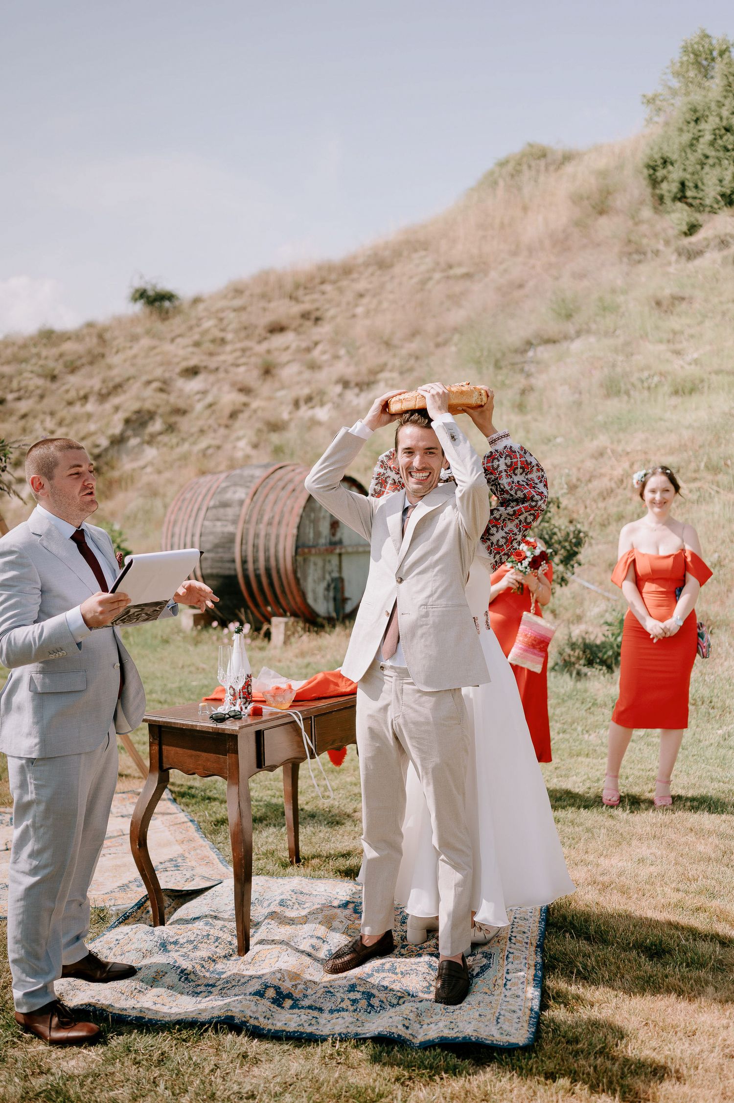 Outdoor wedding celebration with guests in light suits and red accents on a scenic hillside.