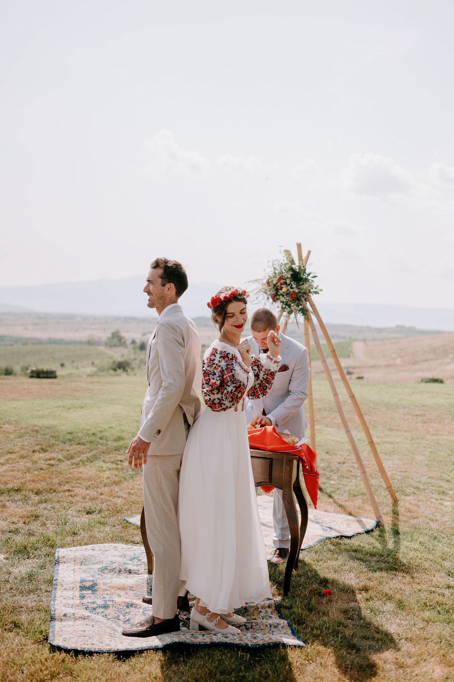 A wedding celebration takes place on a hilltop with wooden easels and decorative rugs.