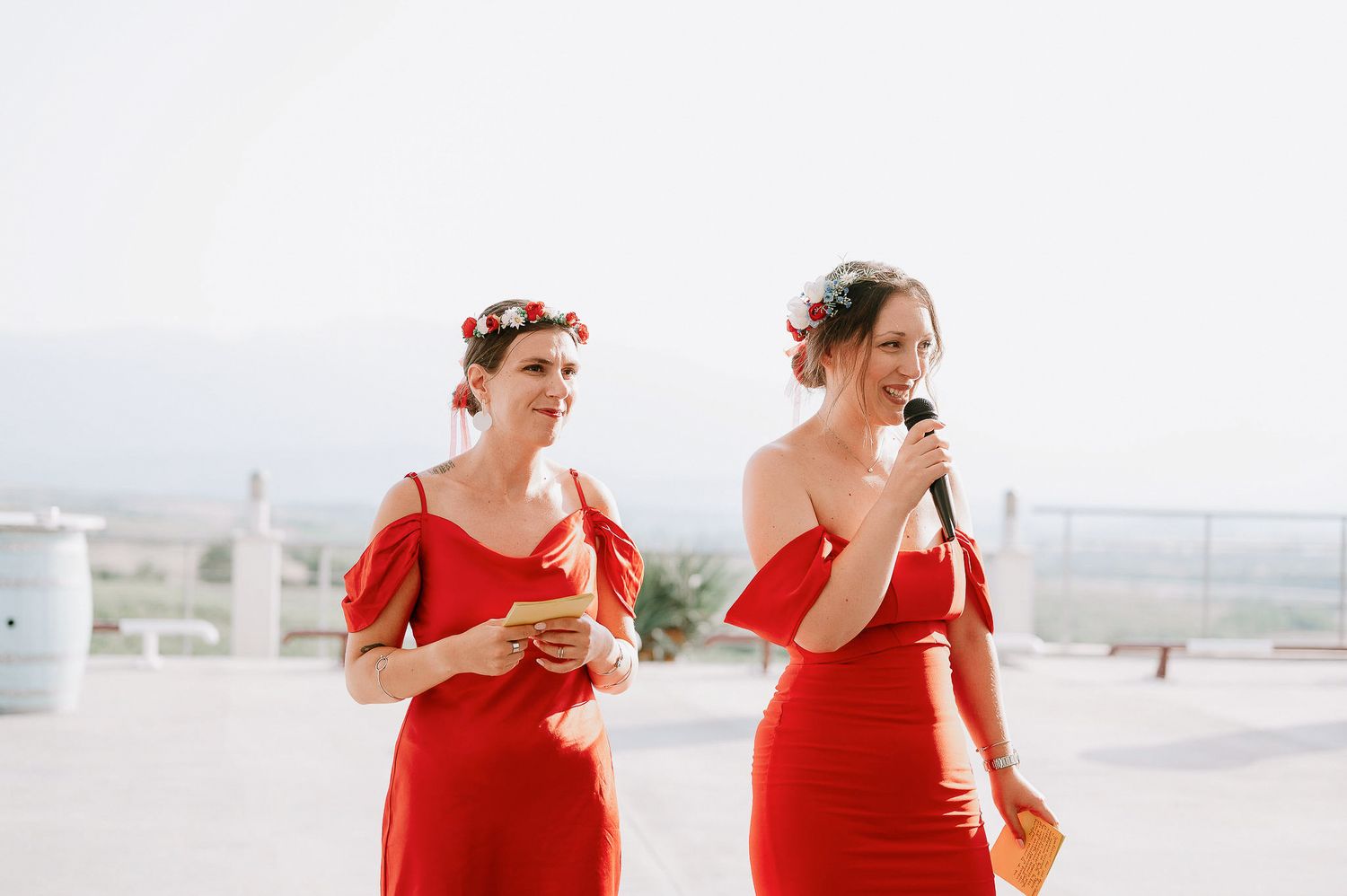 Two people in red dresses stand on a bright outdoor terrace with ocean views.