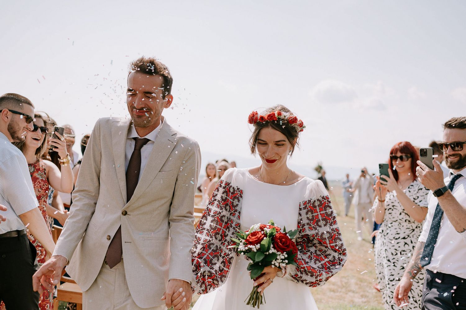 A couple in wedding attire walks hand in hand among guests at an outdoor celebration.