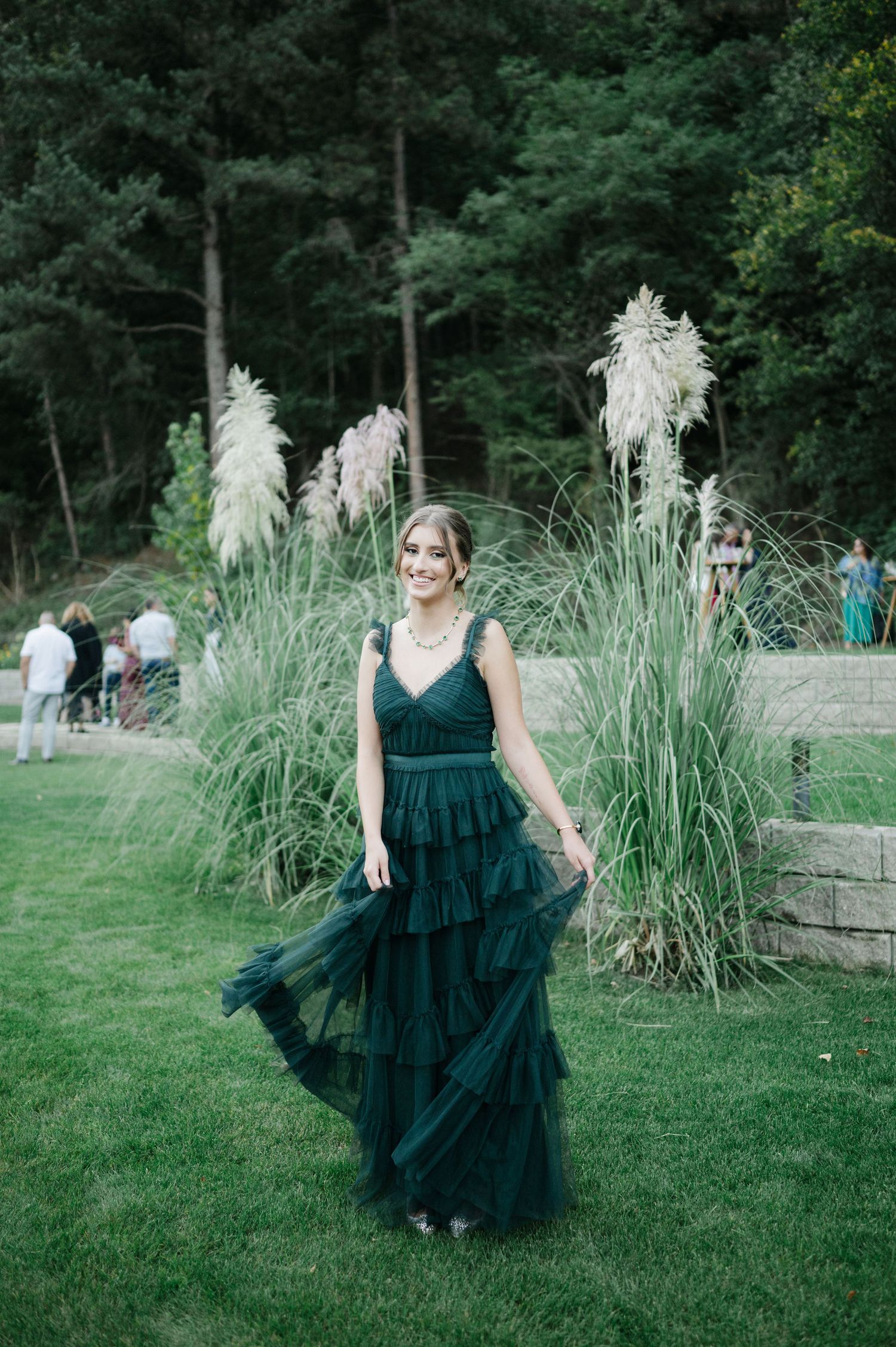 A figure in an emerald green tulle dress dances joyfully on a lawn near decorative grasses.