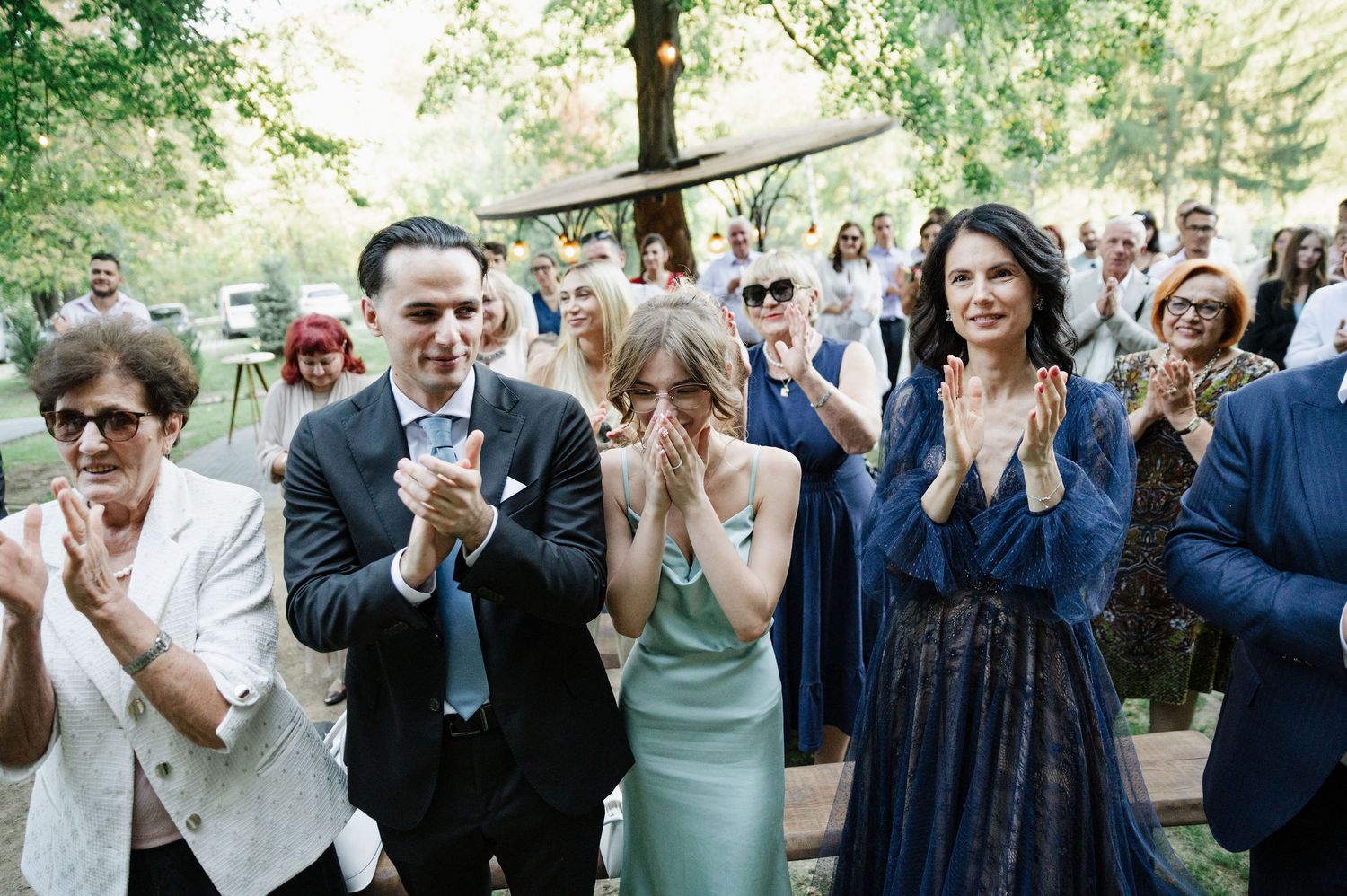 Wedding guests react emotionally during outdoor ceremony in wooded setting.