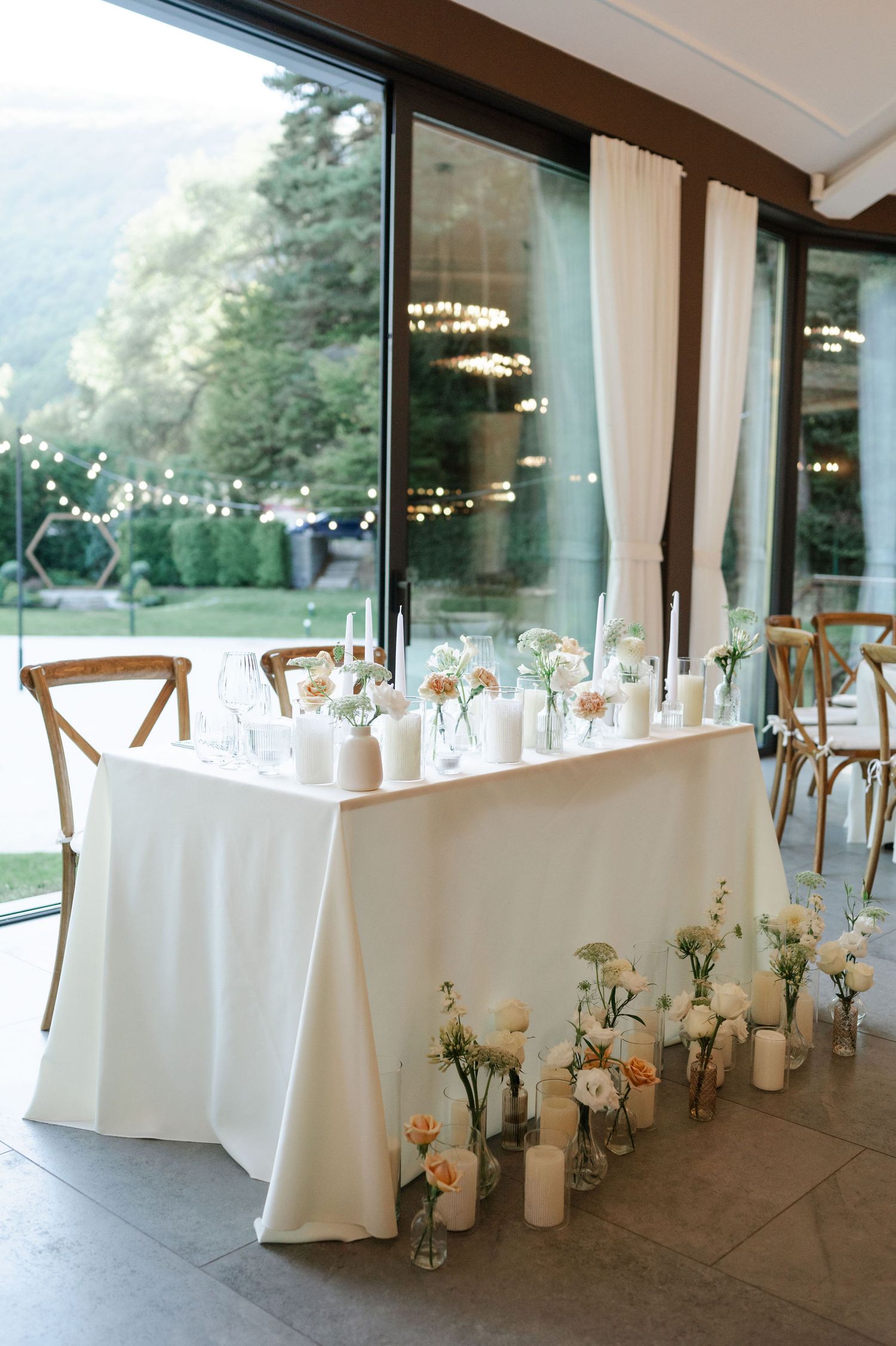 Wedding reception table with white linens and floor vases against large windows overlooking mountain view.